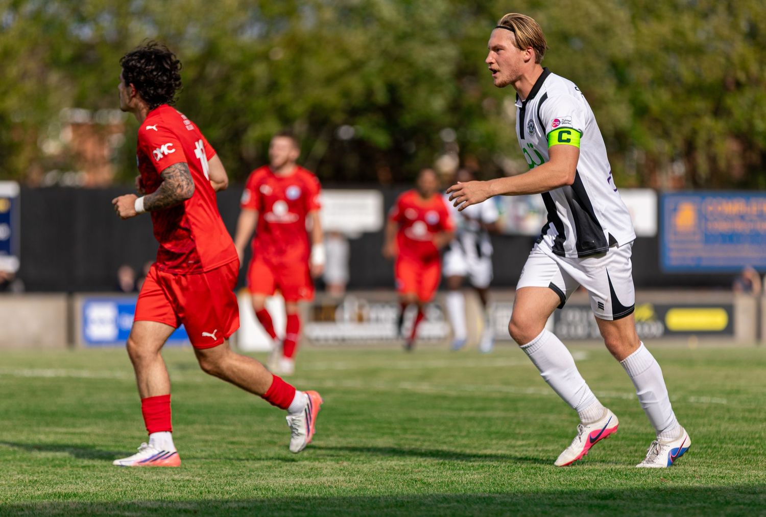 Two soccer players in red and white uniforms compete on a green grass field during a sunny outdoor match.