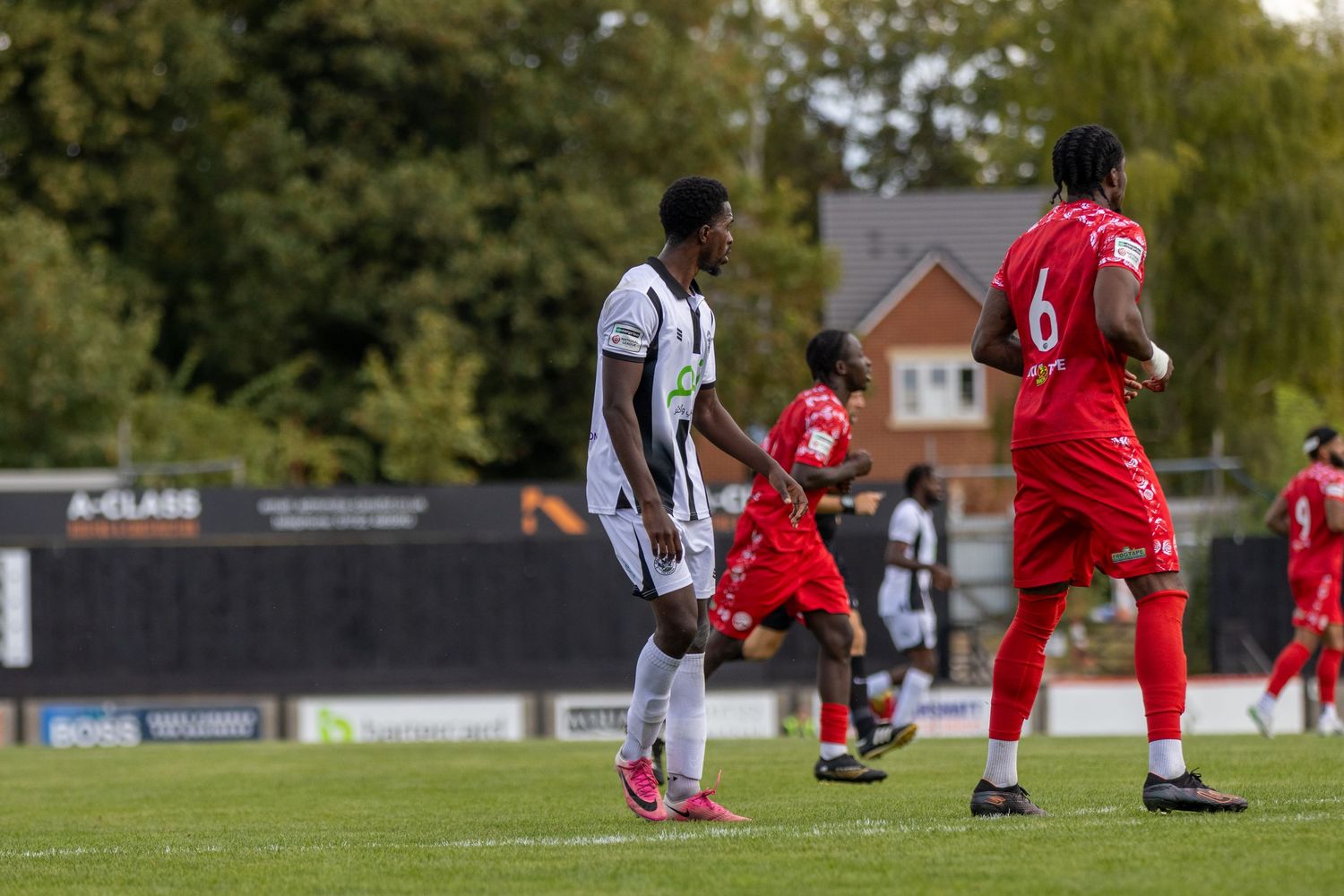 Football players in black-white and red kits compete during an outdoor match on a green pitch with trees in background.