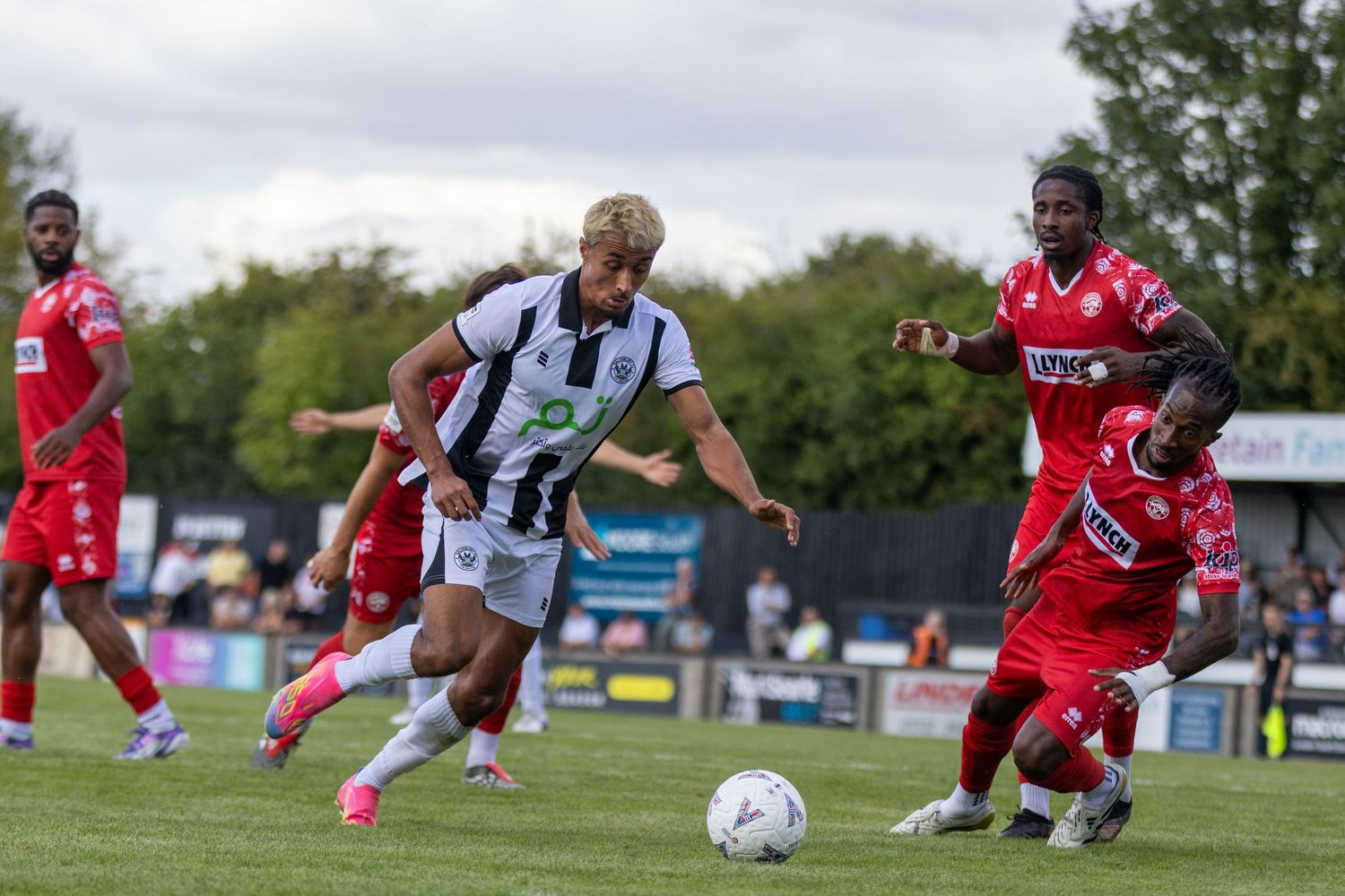A black and white kit player dribbles past red-shirted opponents during a football match.