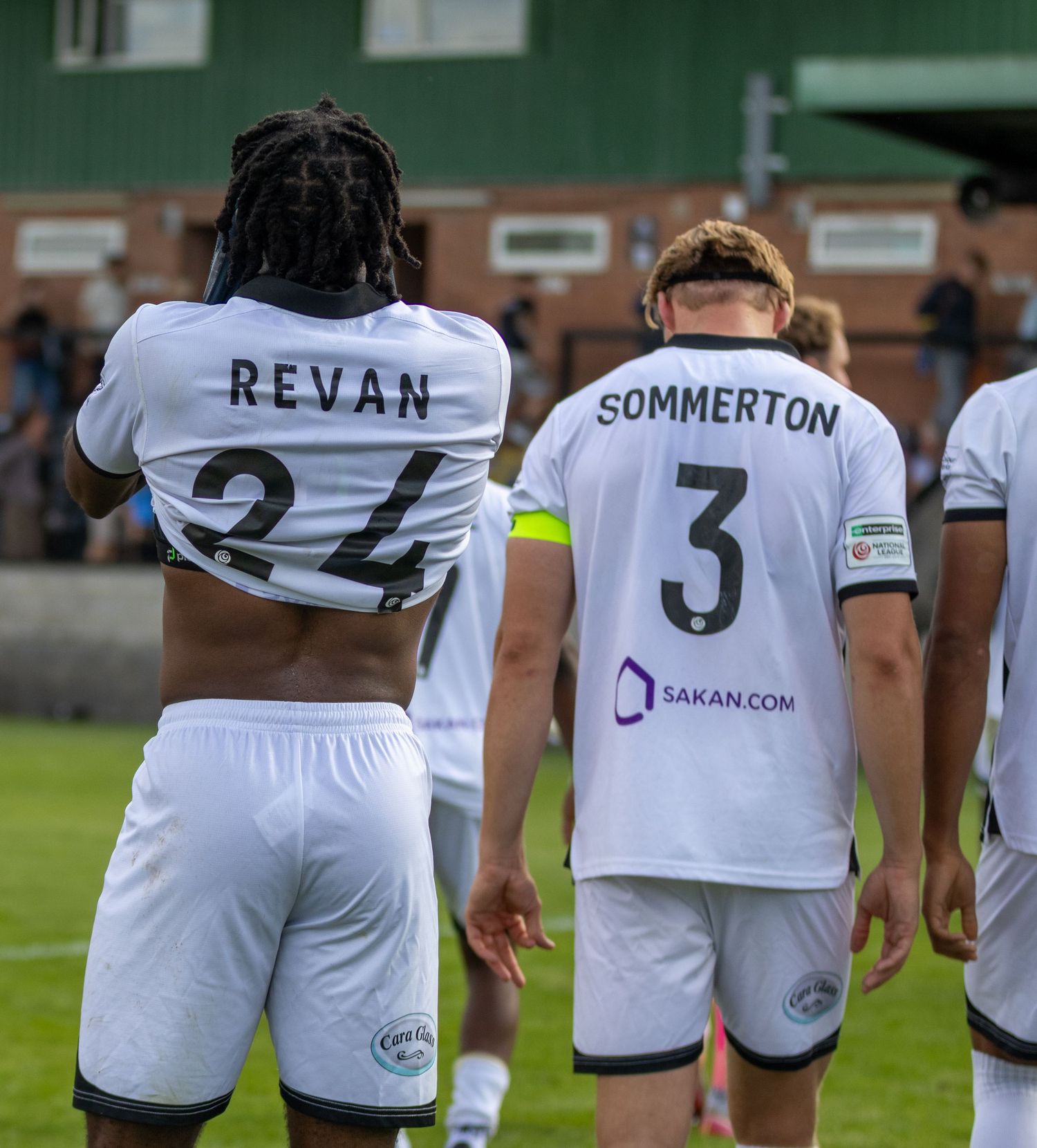 Two football players wearing white jerseys, Revan #24 and Sommerton #3, stand on a green pitch during a match.