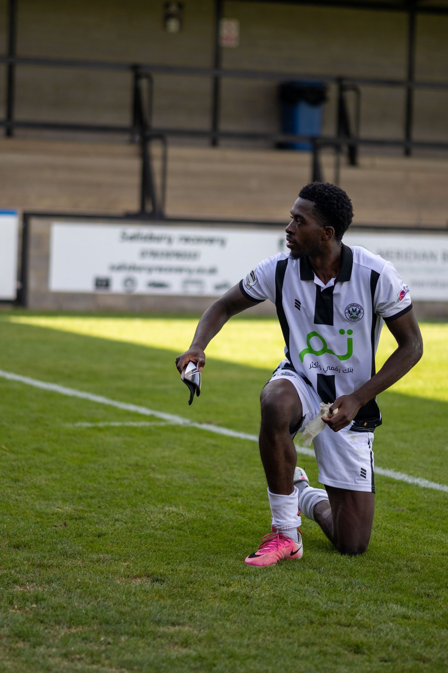 Football player in black and white kit kneeling on grass pitch during a match at a non-league stadium.