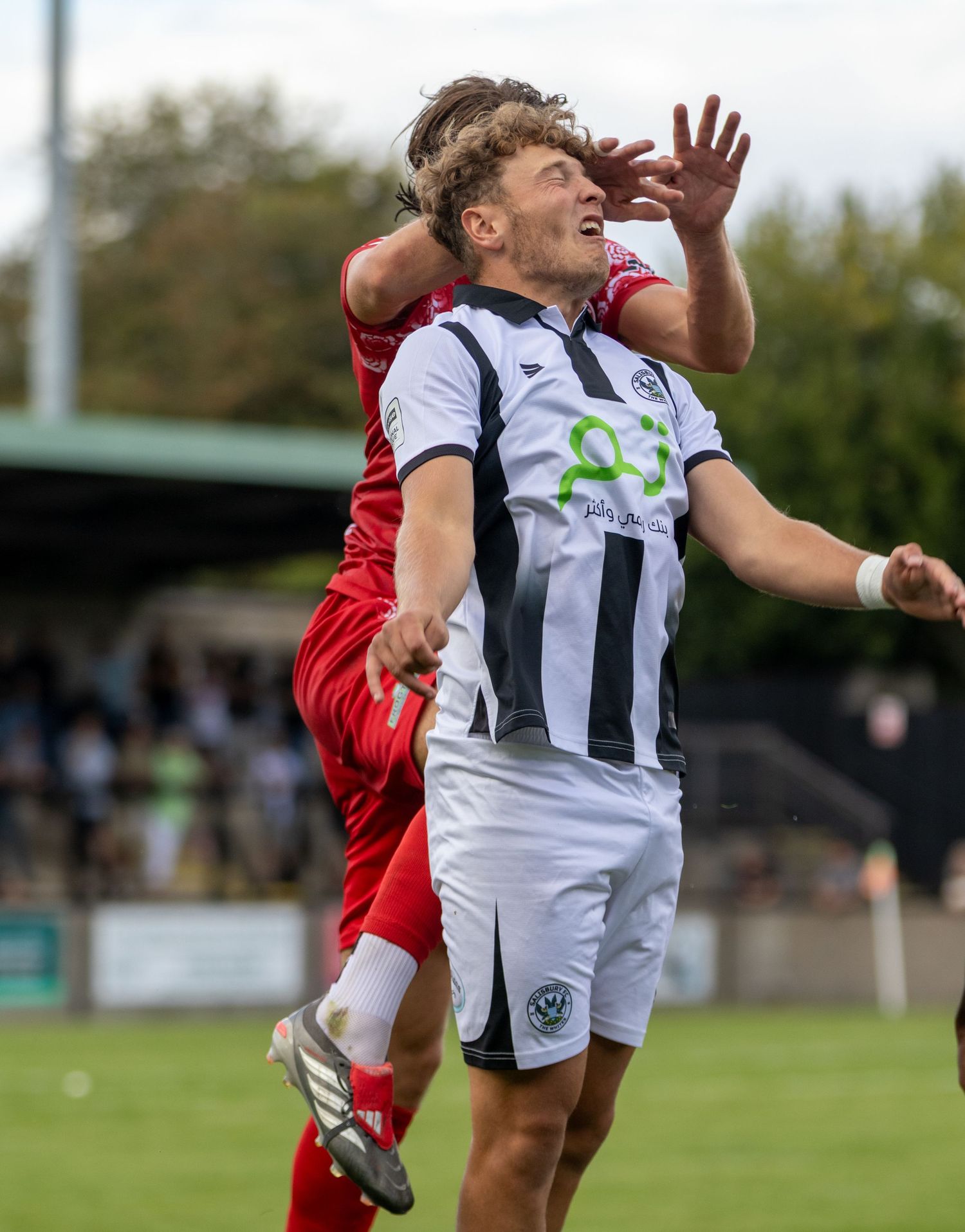 Two soccer players compete for the ball during a match, one in white and black kit, the other in red, on a grass pitch.