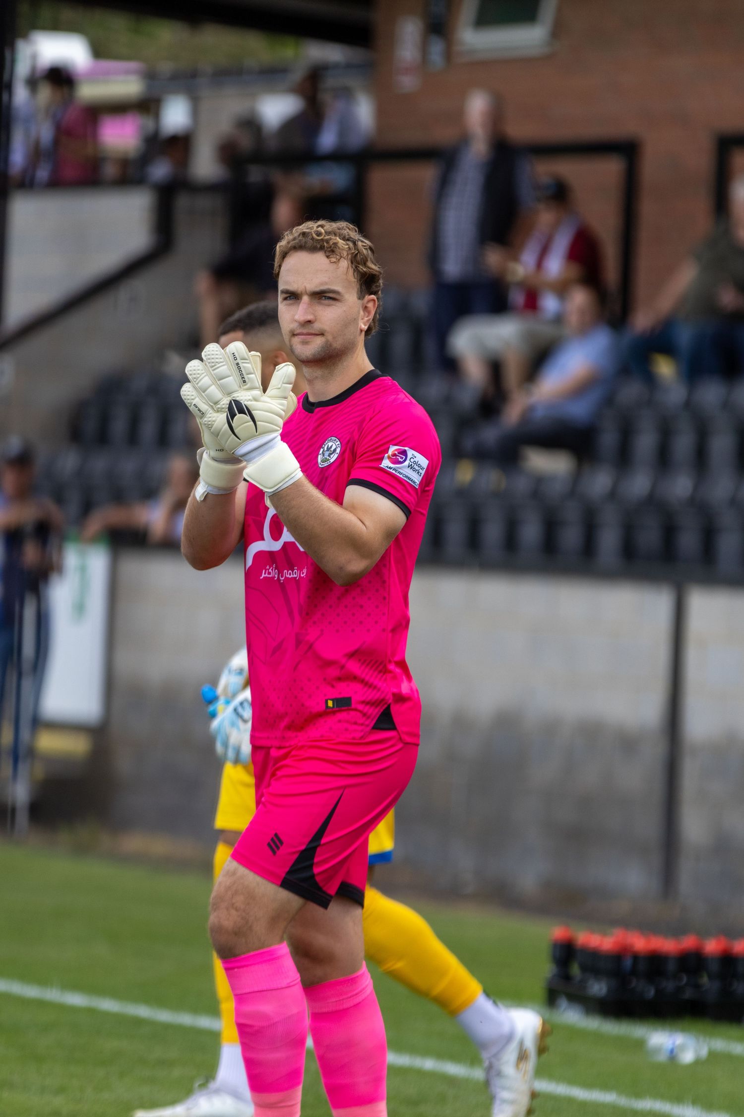A goalkeeper in a bright pink kit claps his gloved hands together on a football pitch during a match.