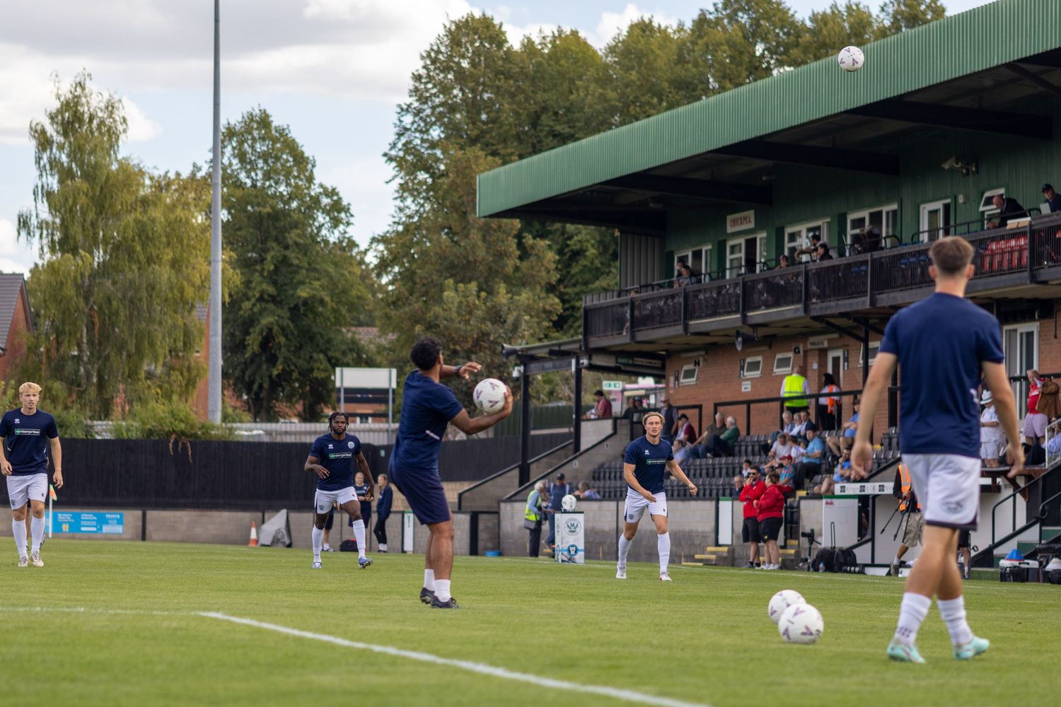 Soccer team in dark blue jerseys warming up with ball drills on a green field at a local stadium.