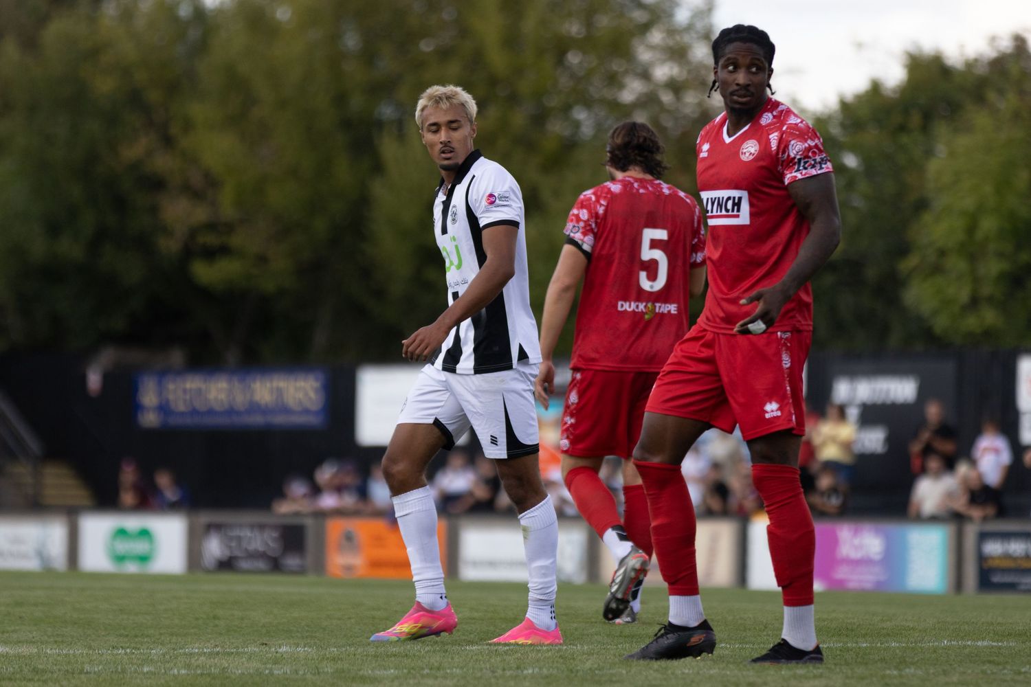 Players in black-white and red kits contest a non-league football match on a sunny afternoon.