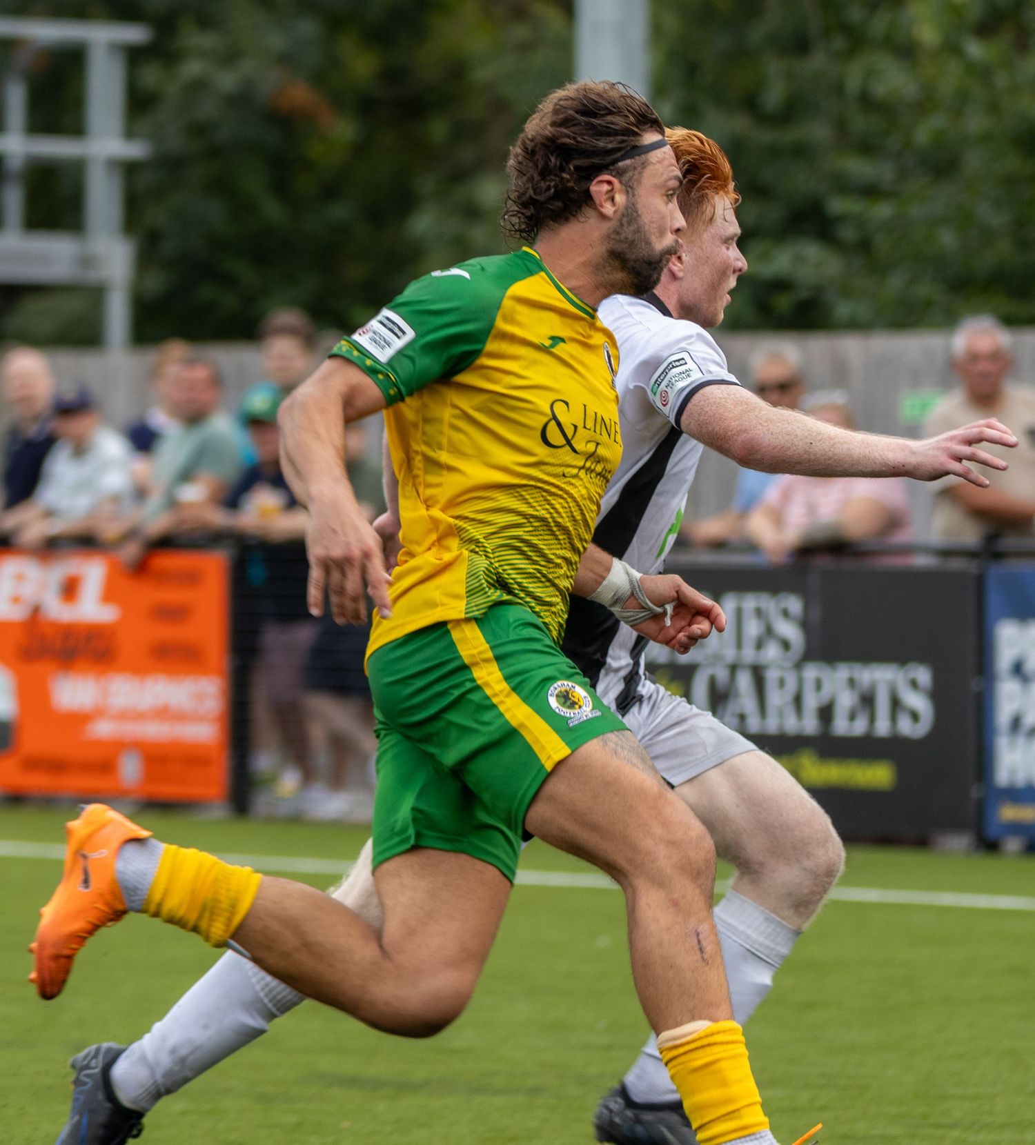 A footballer in yellow-green kit races ahead of a white-black opponent during an outdoor football match.