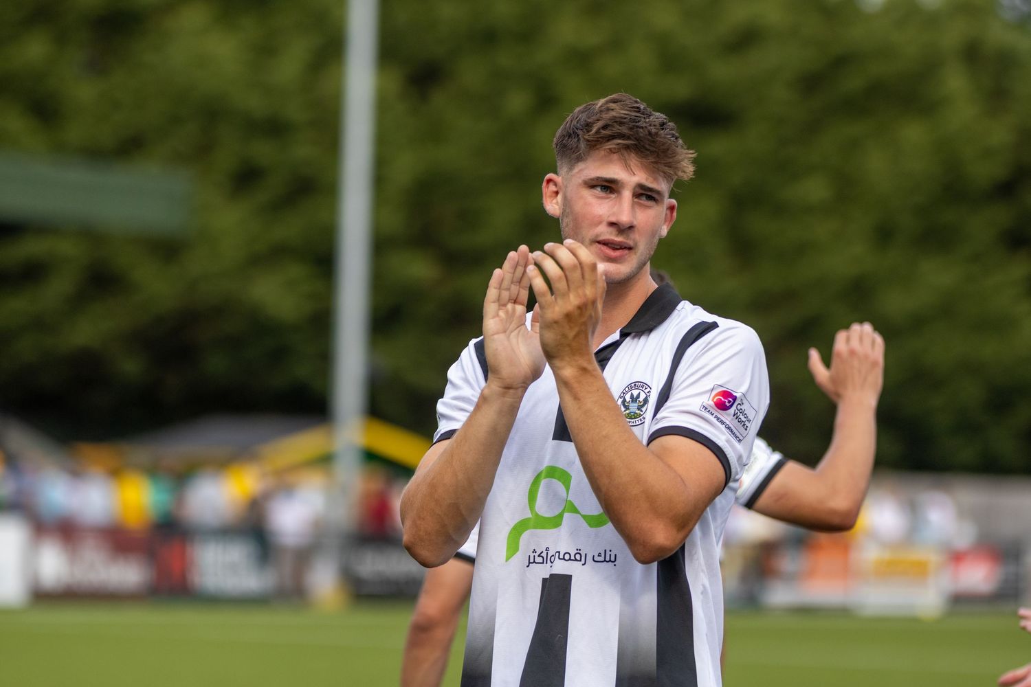 A young male footballer in black and white striped kit applauds supporters after a football match.