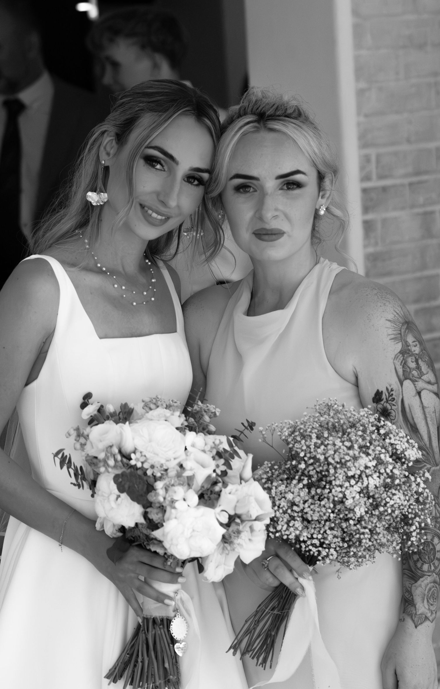 Black and white photo of bride and tattooed bridesmaid in white dresses holding floral bouquets.
