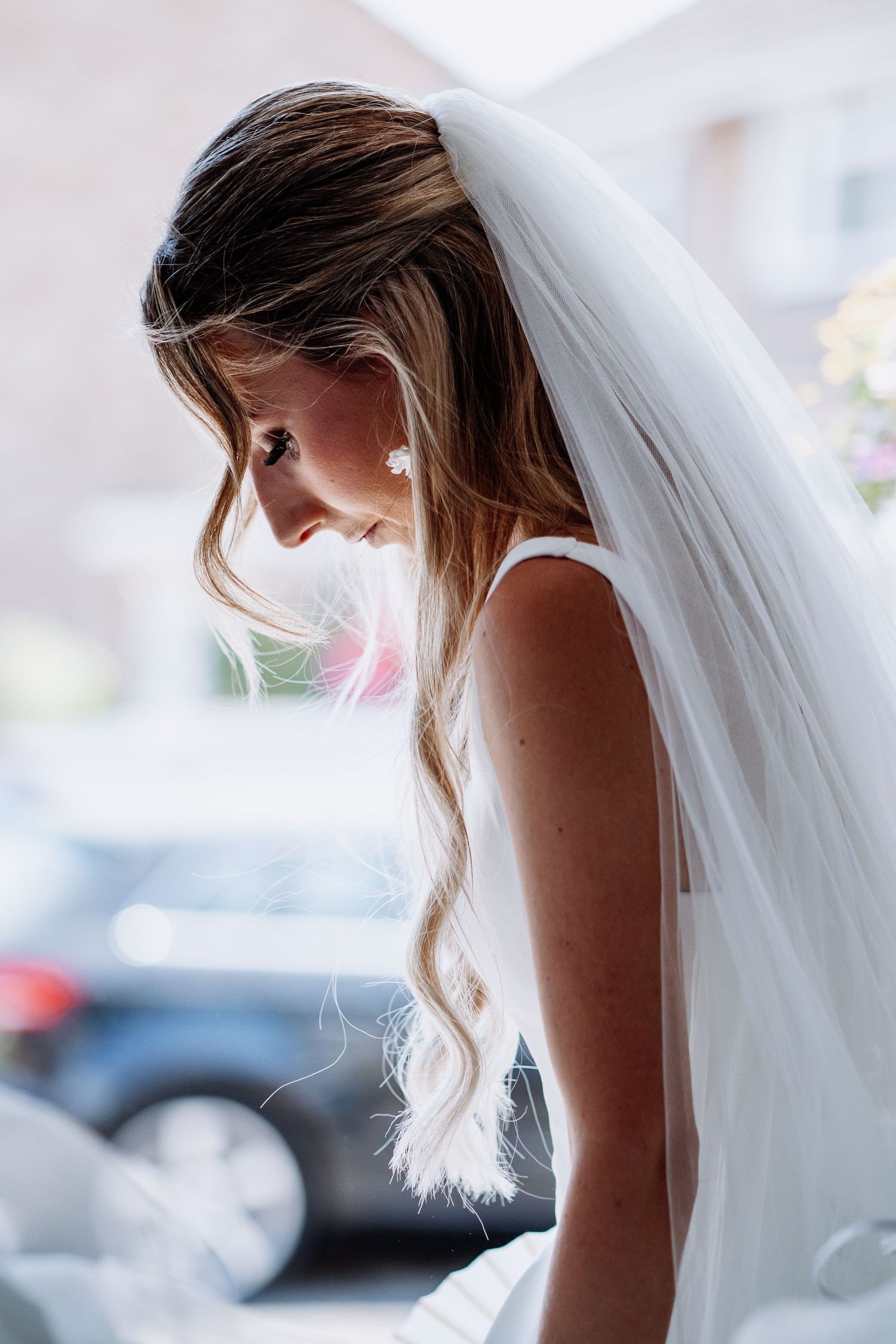 Side profile of bride in white wedding veil with long wavy hair leaning forward in sunlight.