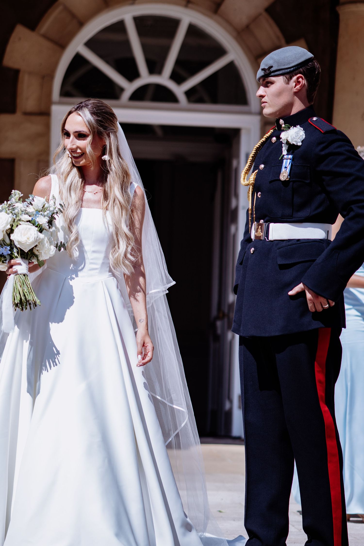 Bride in white off-shoulder wedding gown carries blue floral bouquet beside groom in dark military ceremonial uniform.