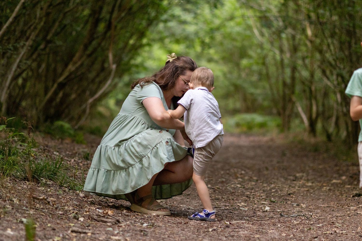 A mother in a green dress crouches down to embrace her smiling toddler son on a forest path.