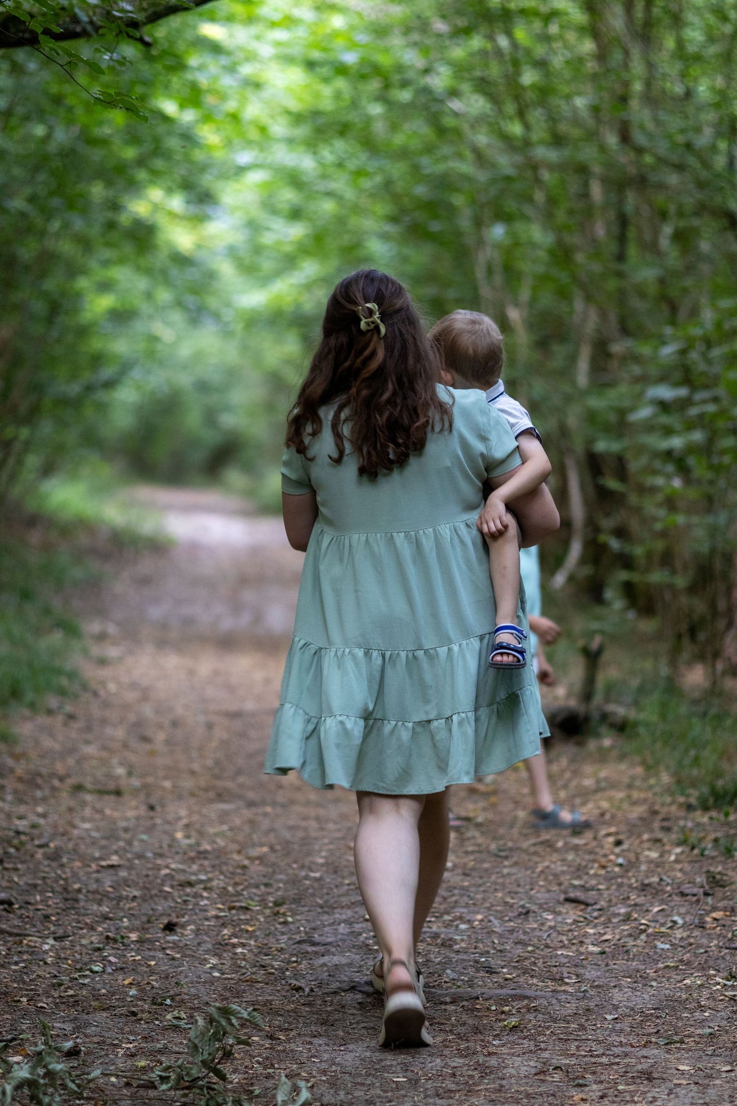 Mother carrying young child away down a sunlit green woodland trail.