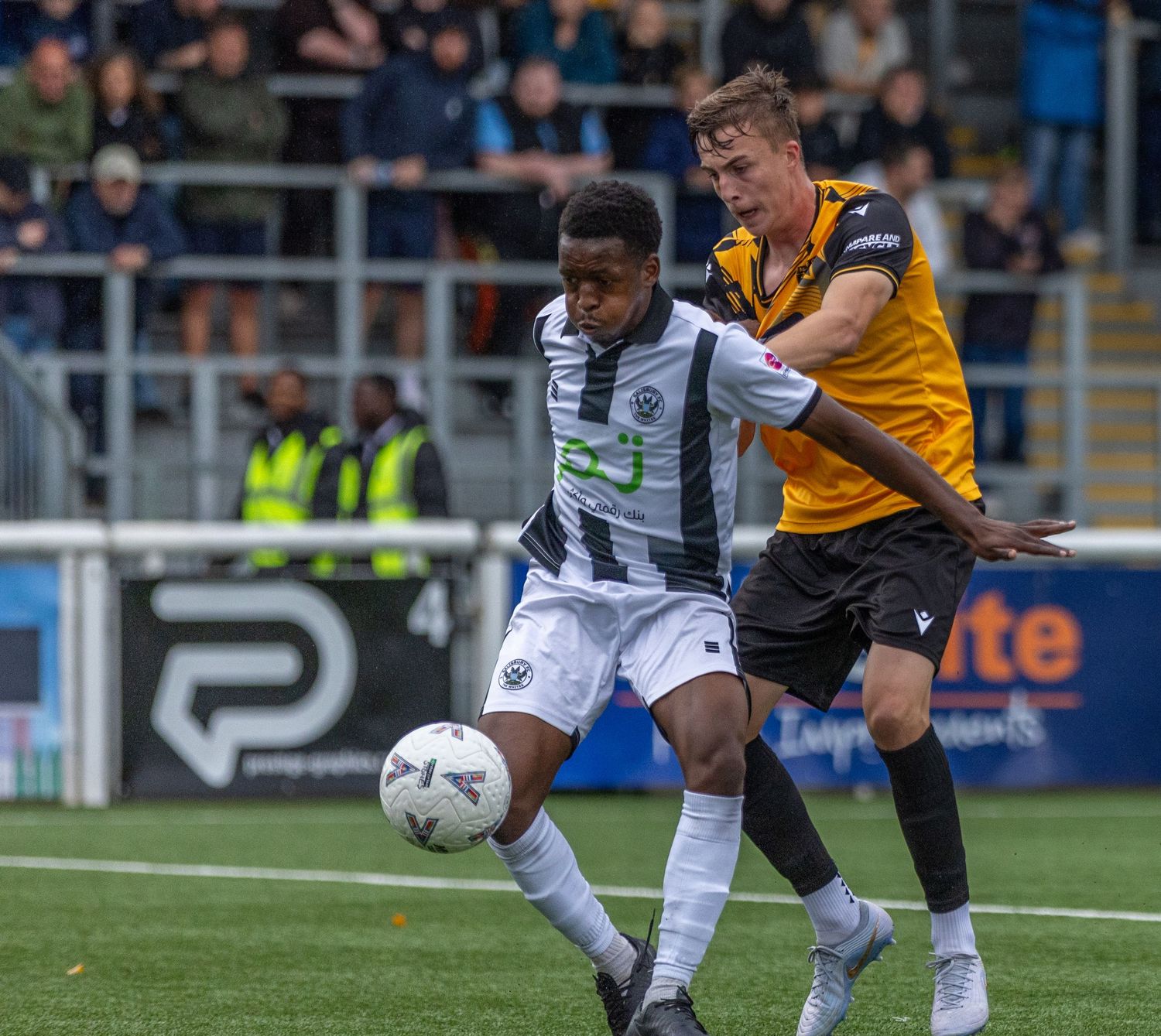 Two soccer players battle for the ball during a competitive match, one in black and white stripes, the other in gold and black.