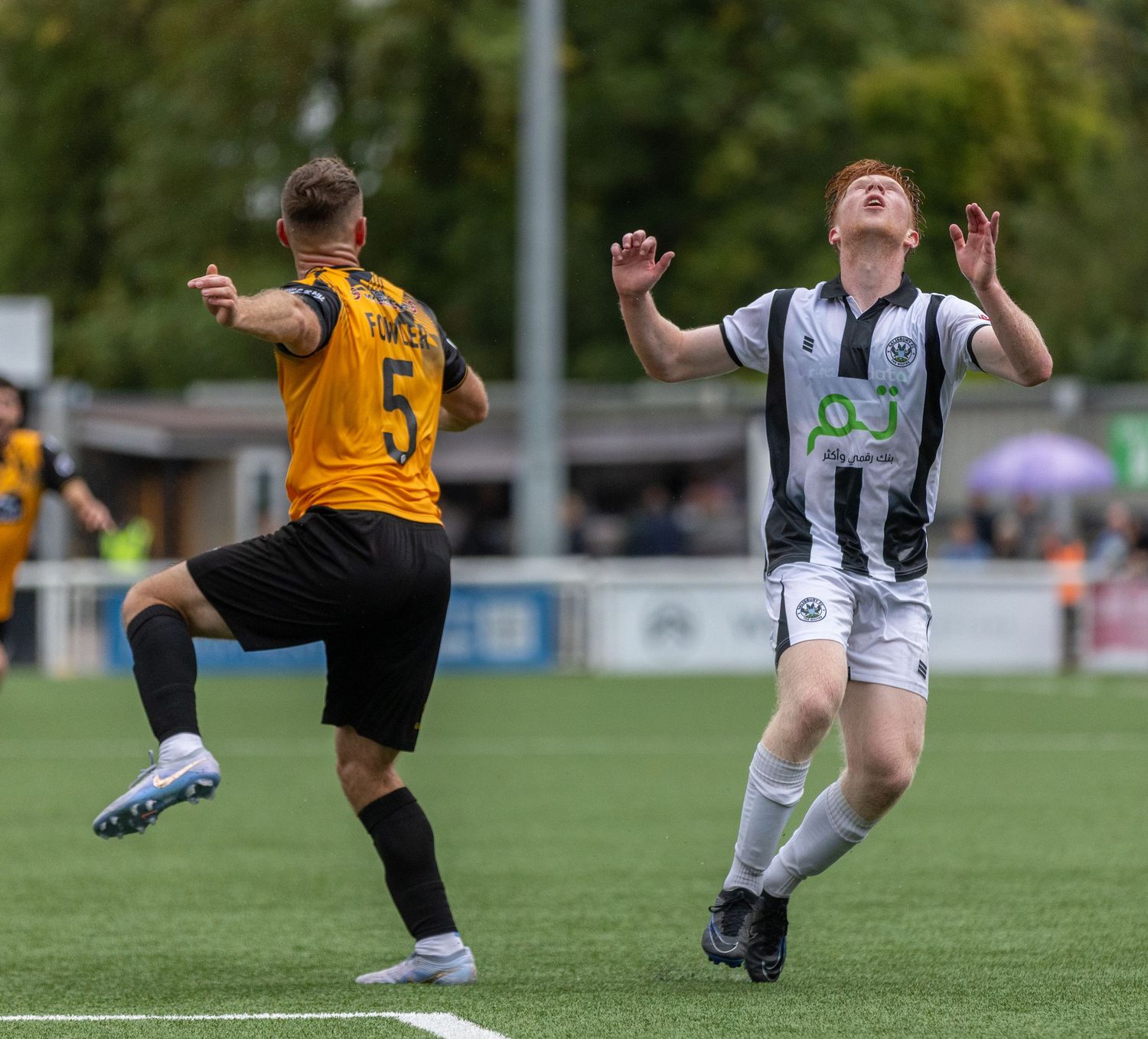 Two soccer players compete for the ball during a match, one in gold and one in black and white striped kit.