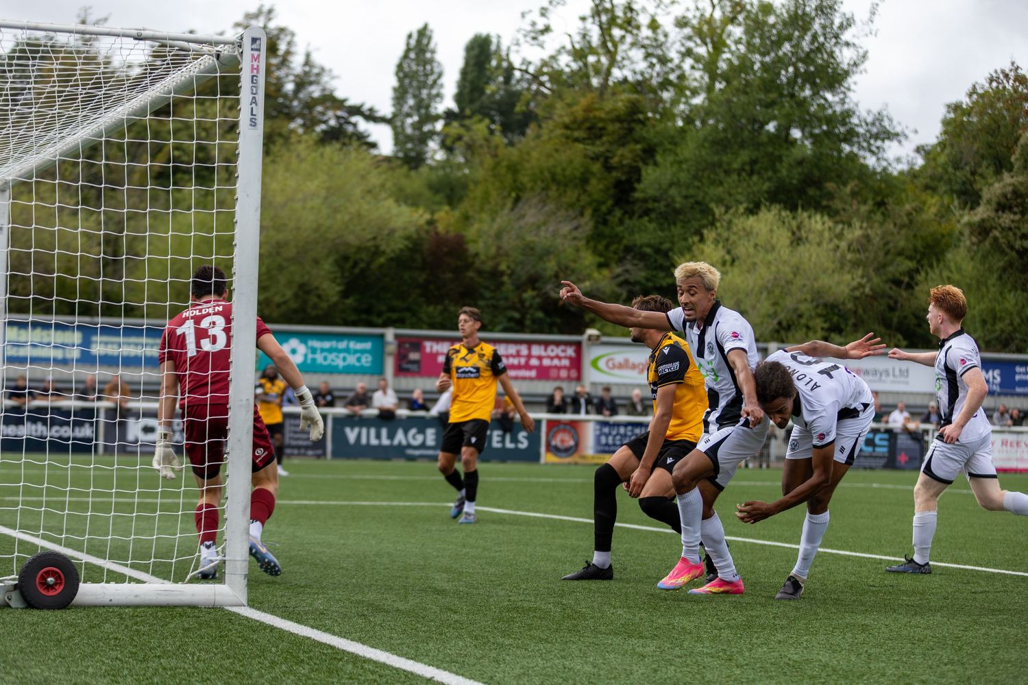 Soccer players compete near the goal during an outdoor match, with one player pointing while others challenge for the ball.
