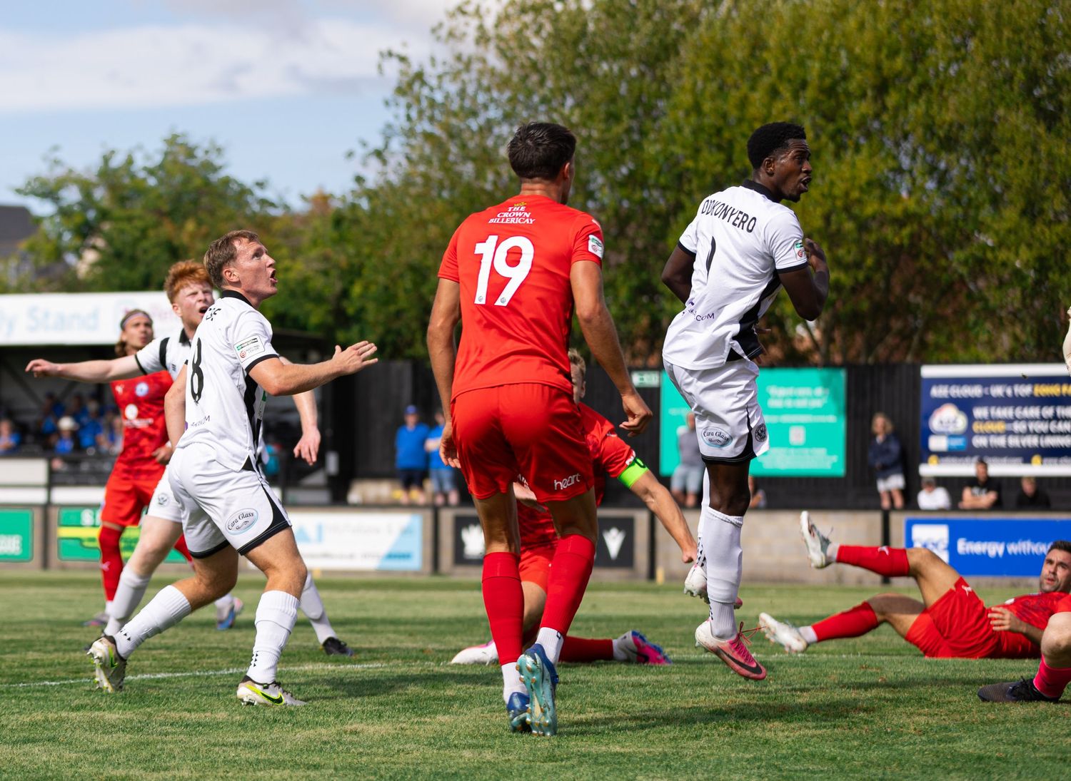 Football players in red and white kits compete during an outdoor soccer match on a green pitch under cloudy skies.