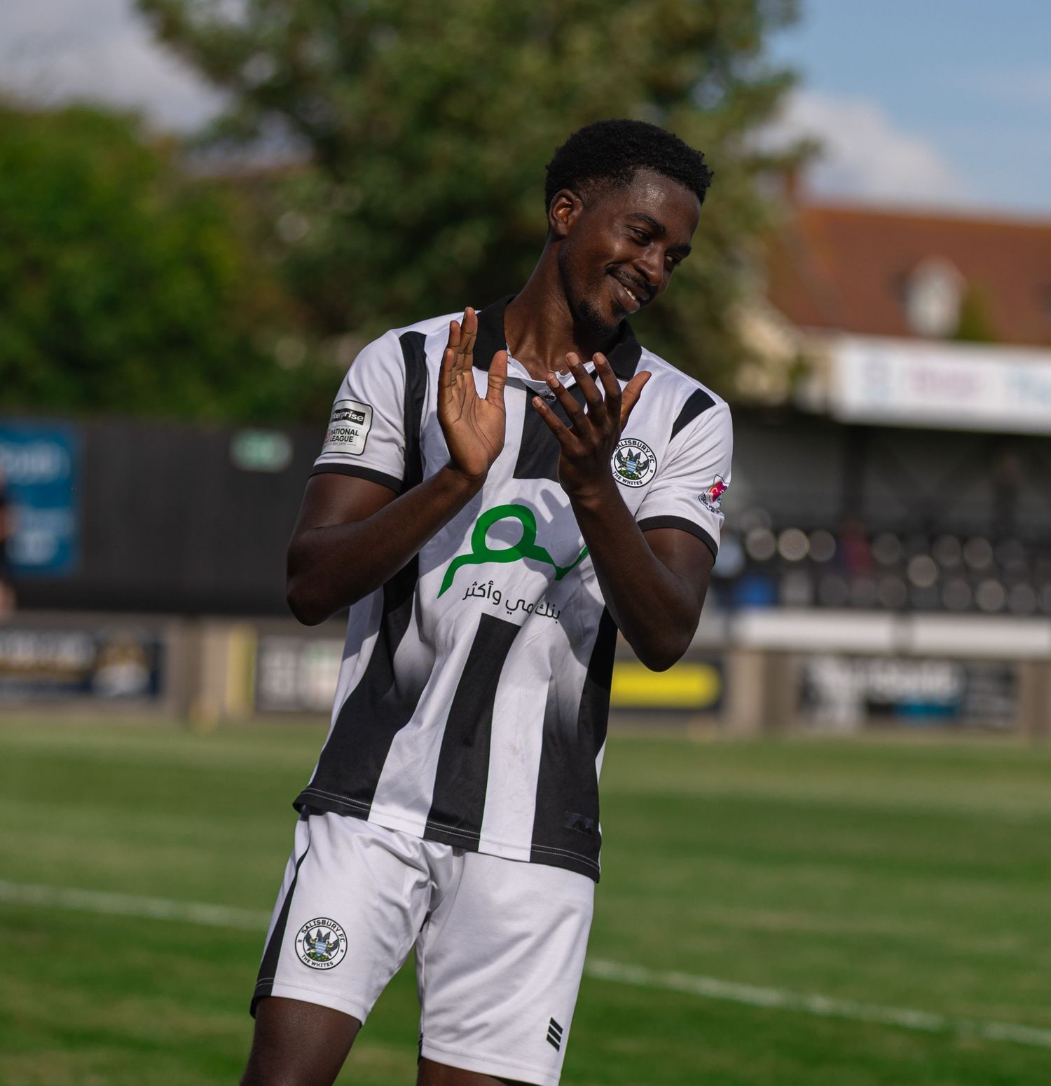 A footballer in black and white striped kit claps hands on a sunny match day at a grass pitch stadium.