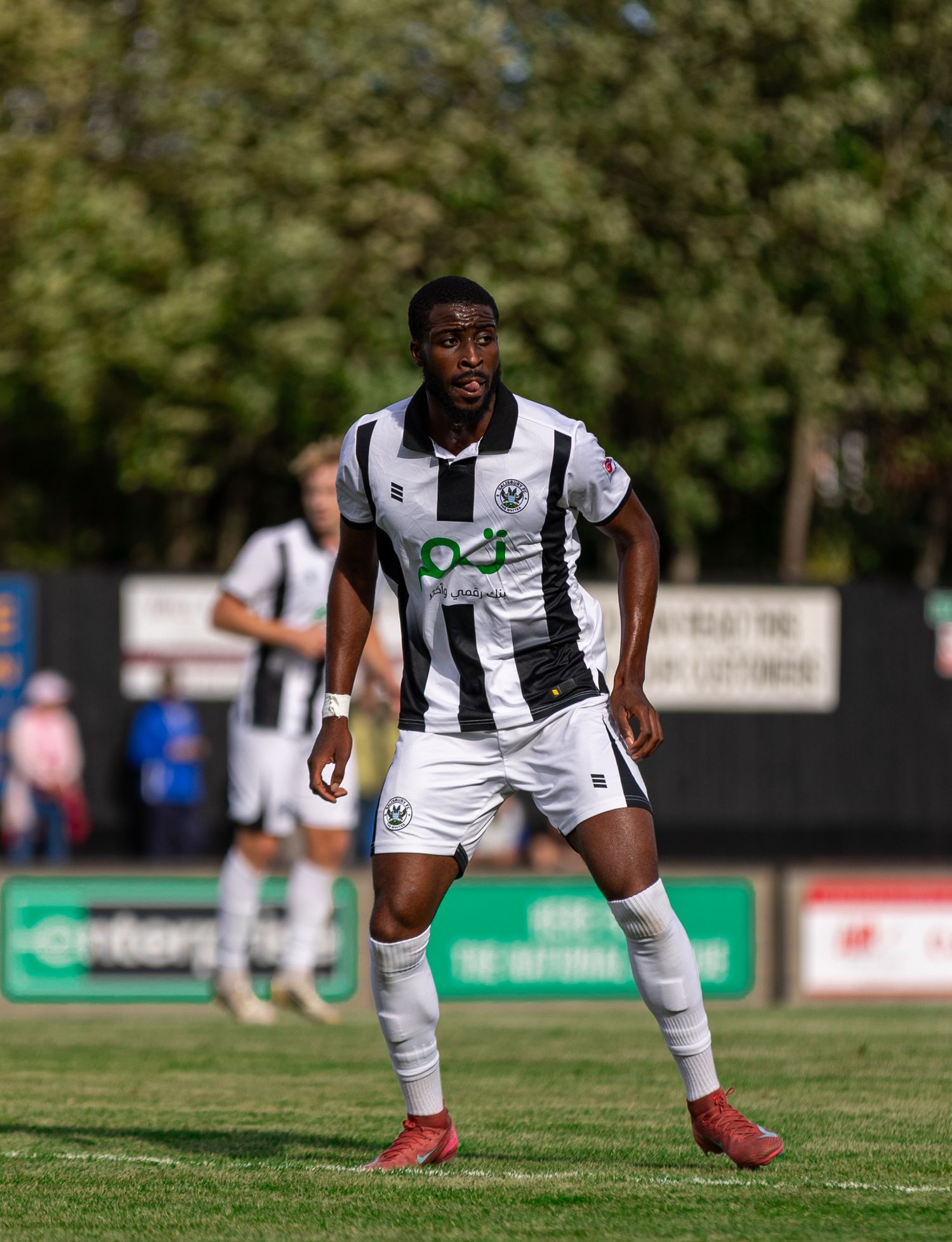 A footballer in black and white kit stands on a grass pitch during a sunny match day.