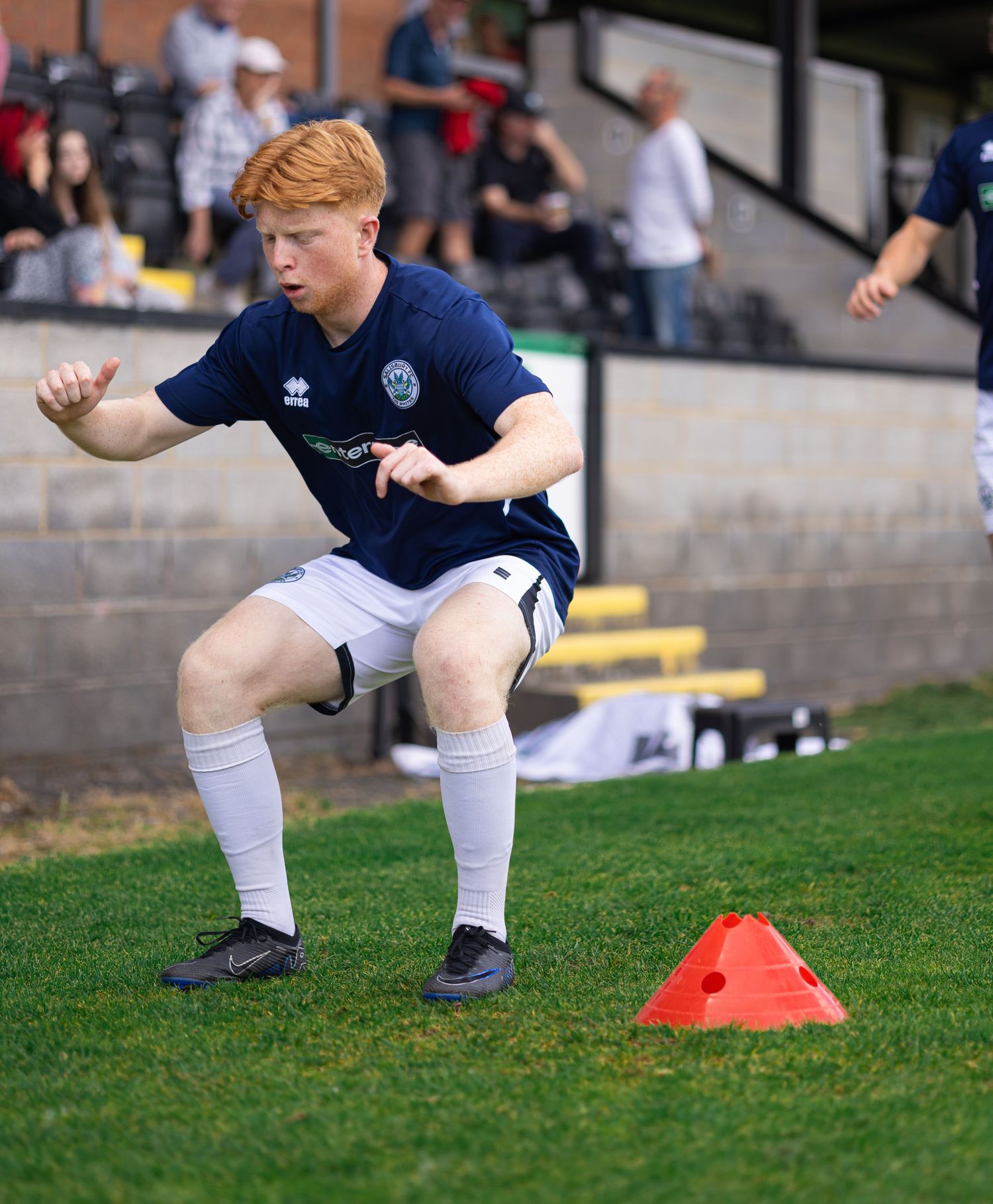 Young red-haired soccer player in navy kit performs agility drill around orange cone on green pitch.
