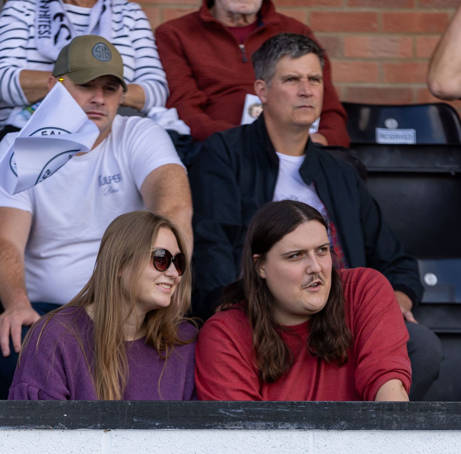 Spectators watching a sporting event from the stands, including a young woman in purple and a man in red.