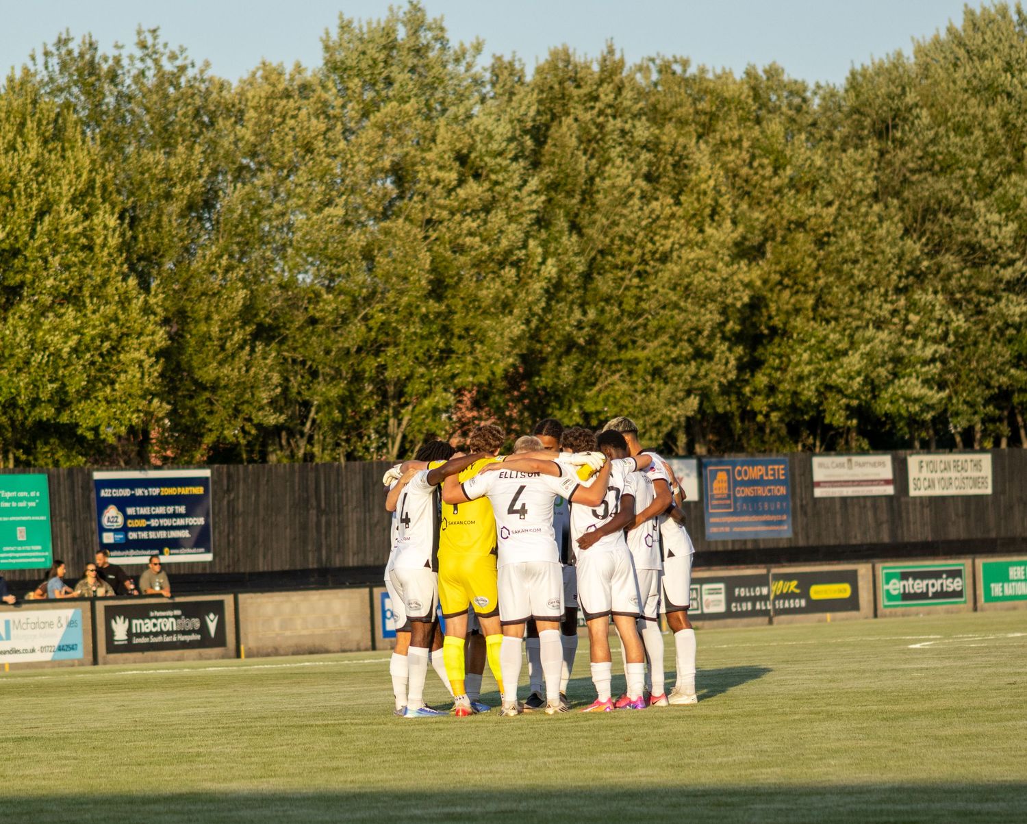 A football team huddles together on the pitch surrounded by green trees before a match begins.