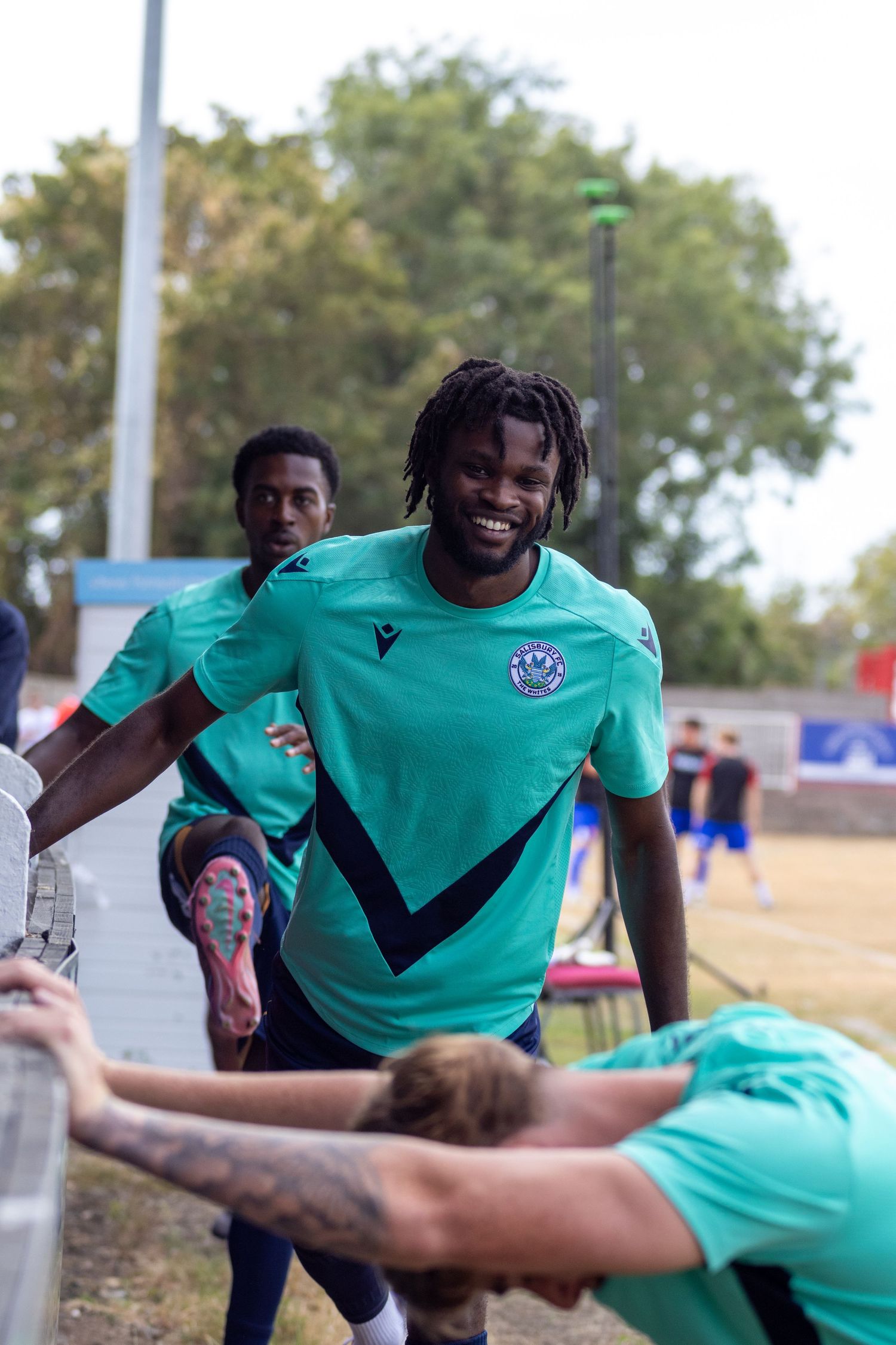 Happy footballer in teal Macron kit laughing while stretching with teammates pitchside.