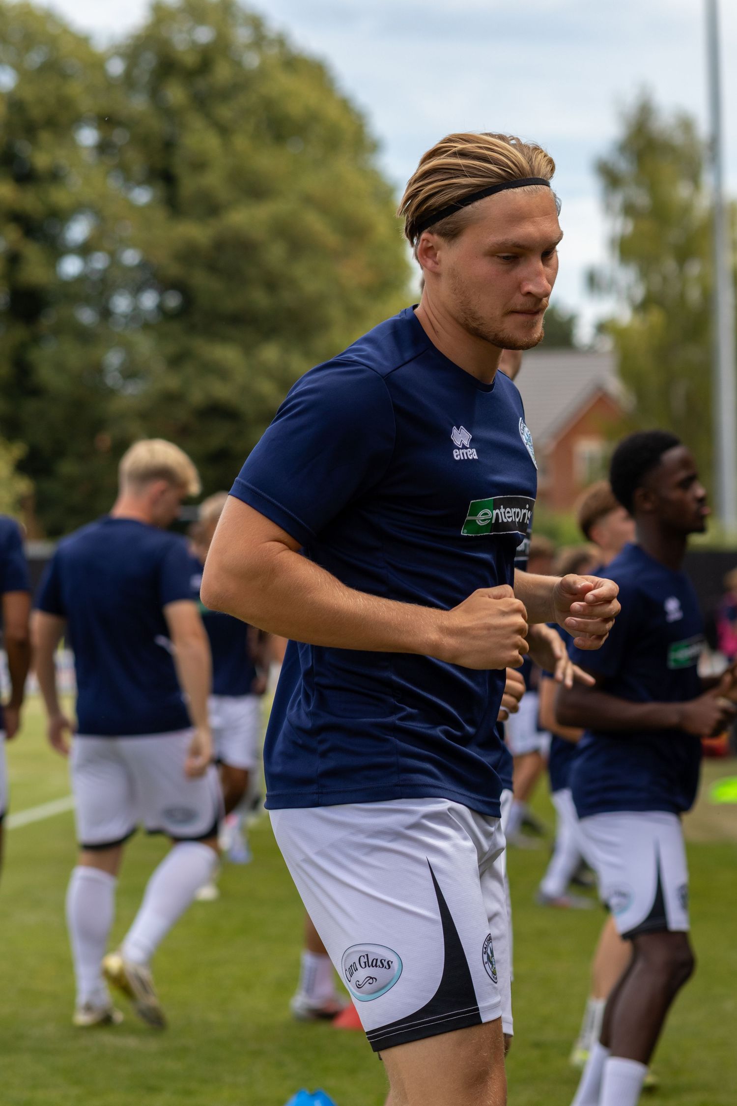 Blonde footballer with hair band in navy Enterprise kit running during outdoor pre-match warmup drill.