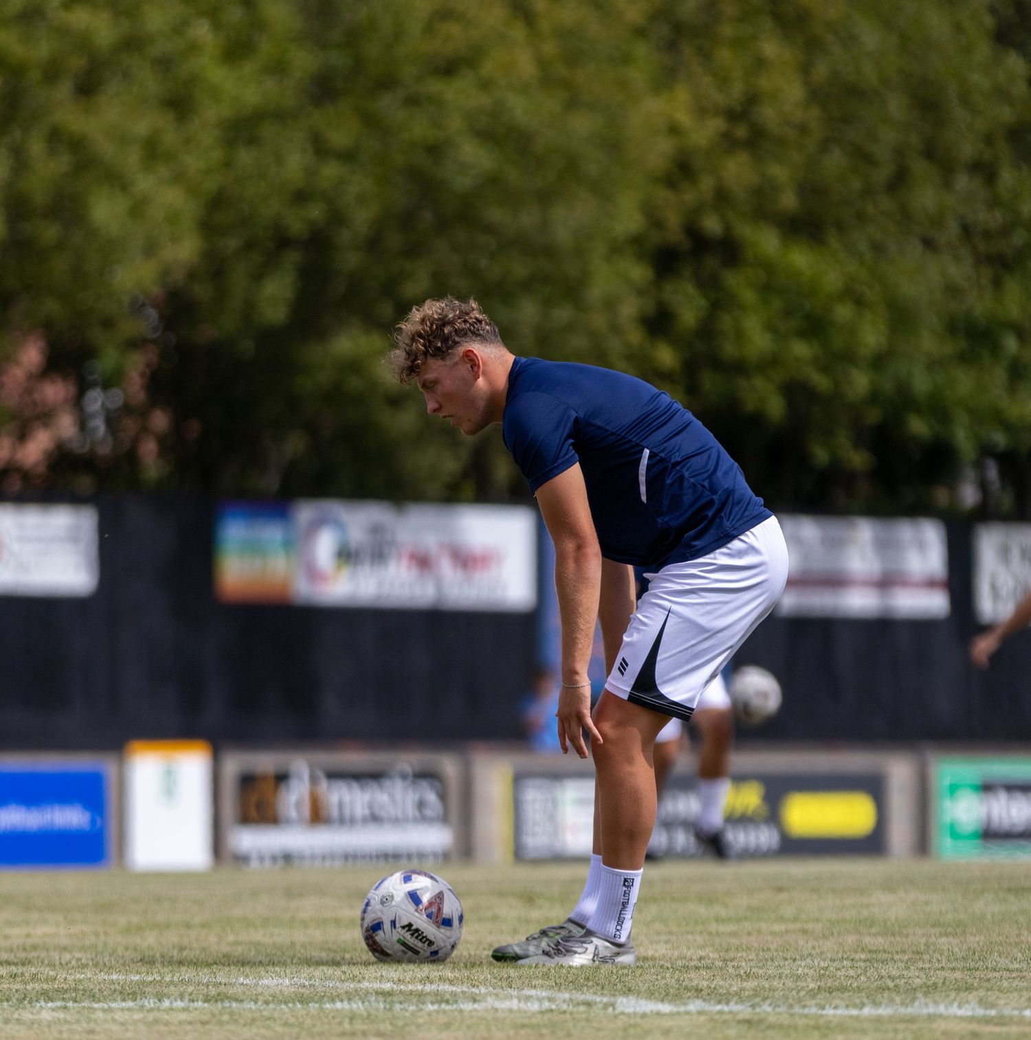 A young footballer in a navy blue kit bends to control a soccer ball on a grass pitch during warm-up.