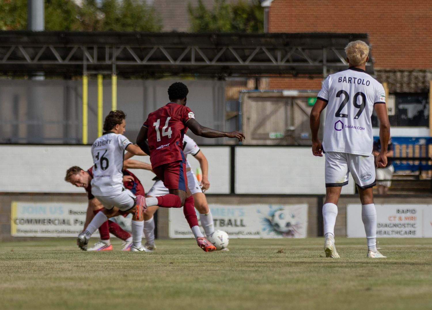 Player number 14 in red shoots during a competitive non-league football match on a sunny day.