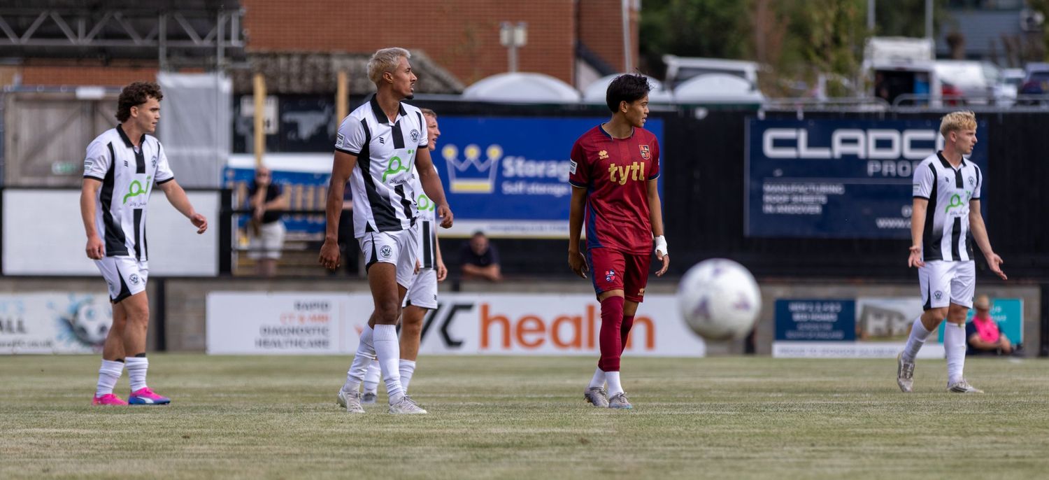 Football players in black and white strip walk on pitch during a match with advertising boards behind.