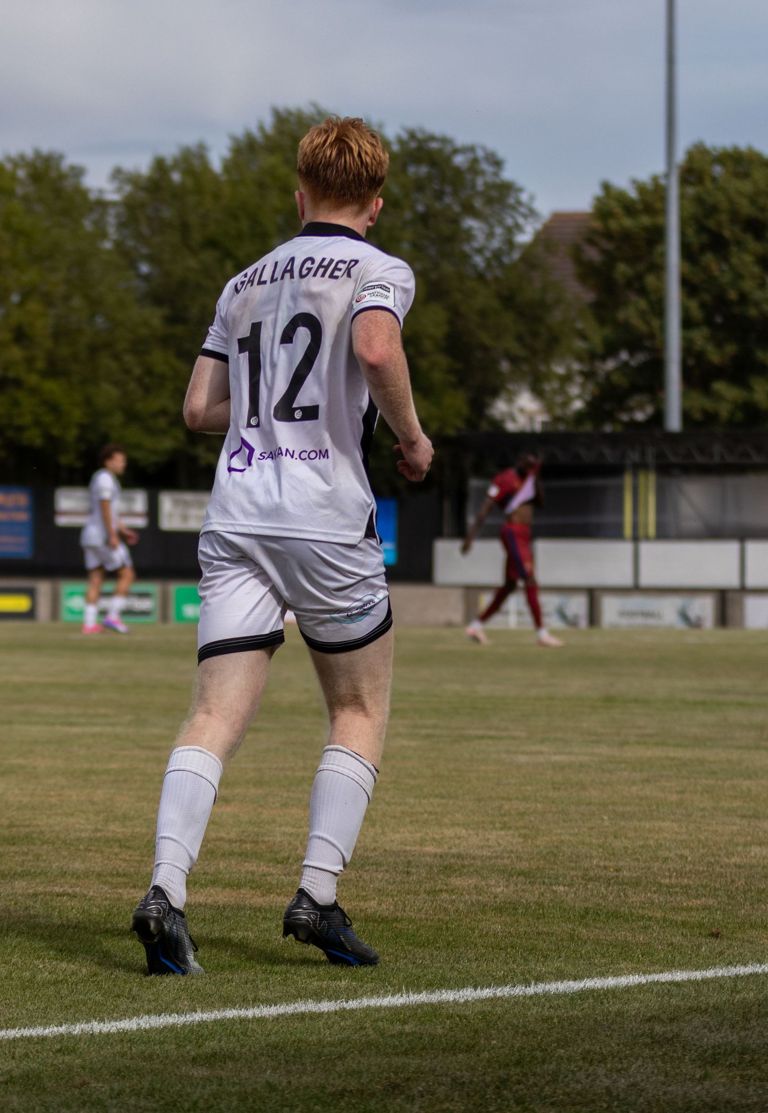 Soccer player Gallagher wearing number 12 white jersey walks on green pitch during a sunny match.