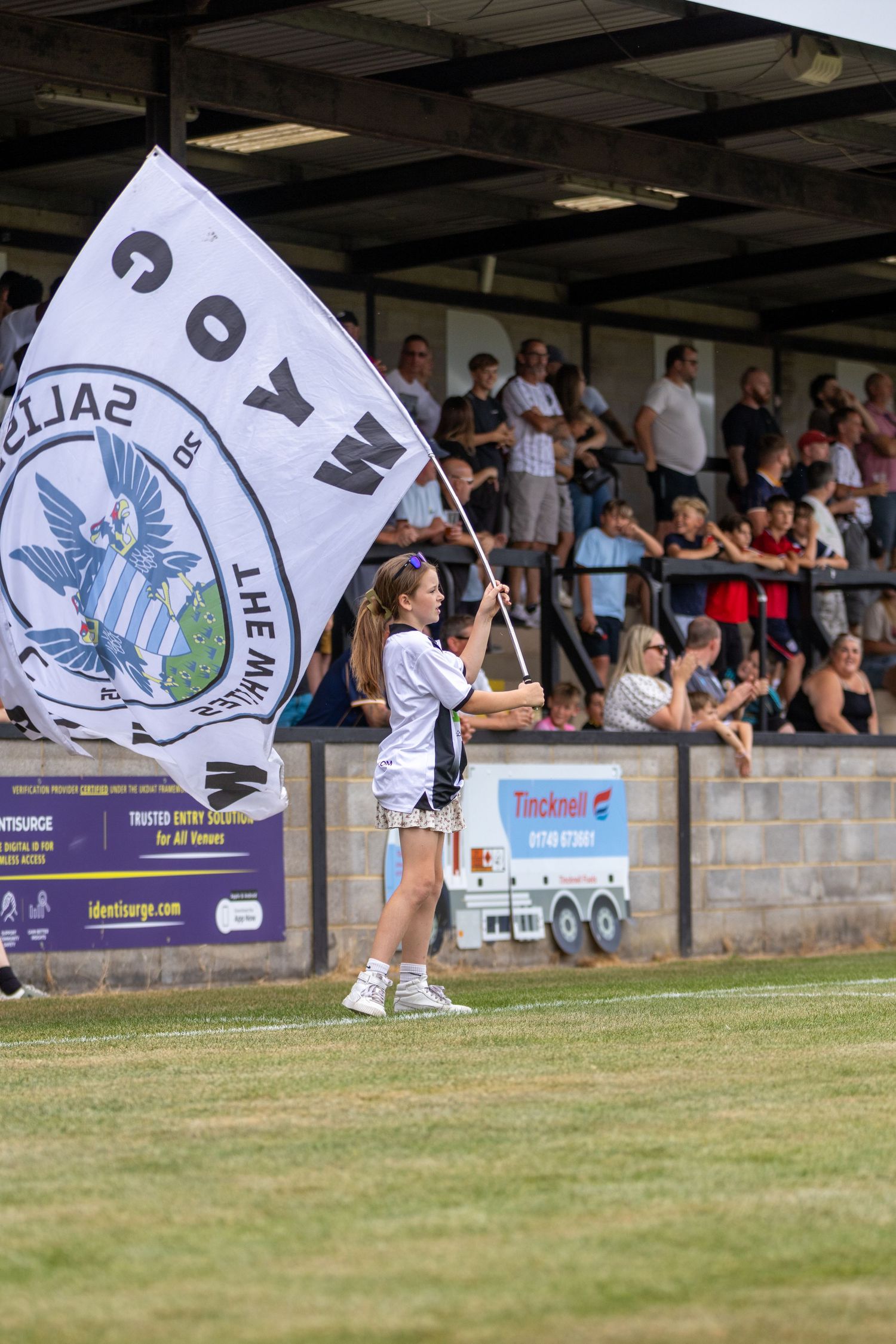 A girl waves a large black and white football club flag on the pitch in front of spectators.
