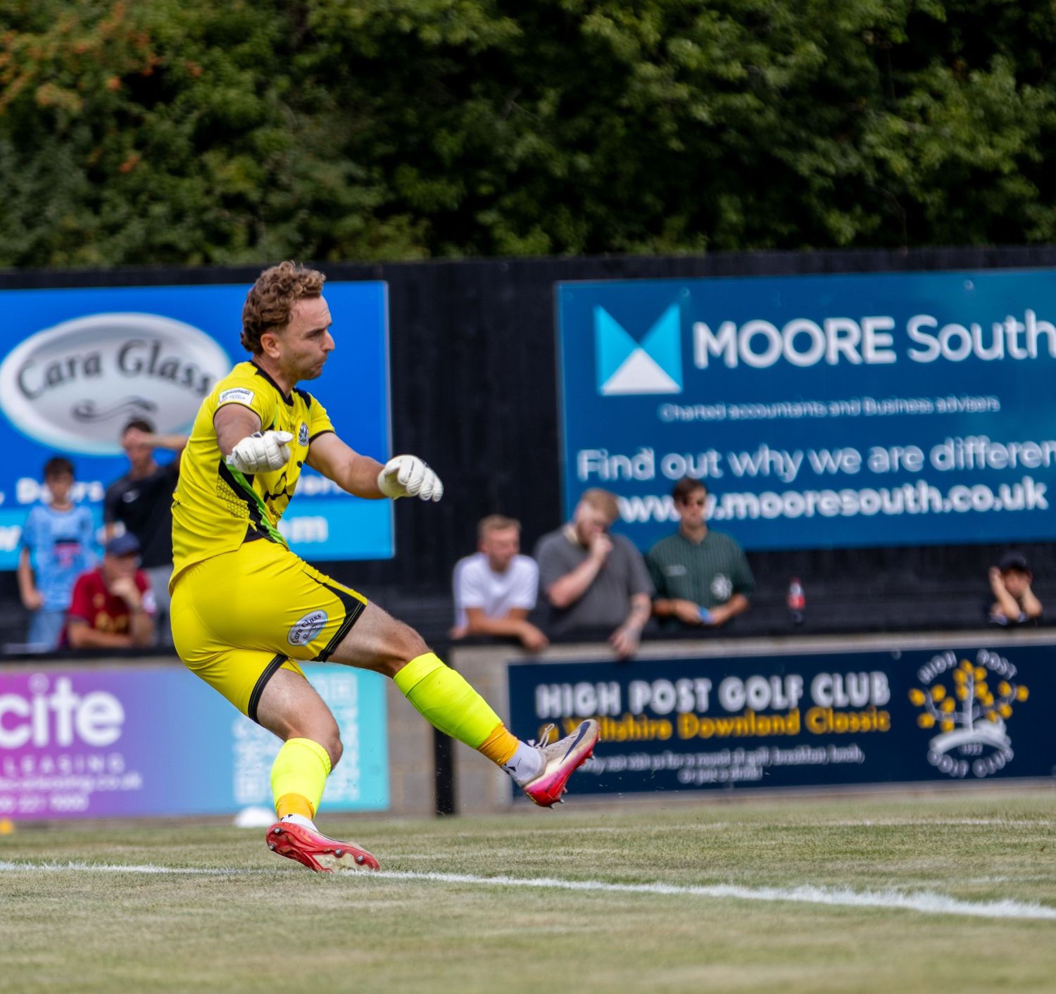 Goalkeeper in yellow kit takes goal kick during sunny non-league football match with advertising boards.