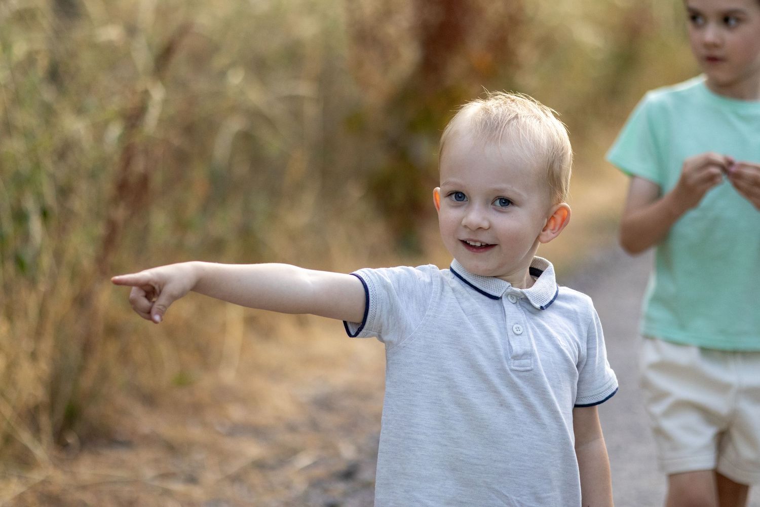 A smiling blonde toddler points excitedly while another child stands behind him on a nature path.
