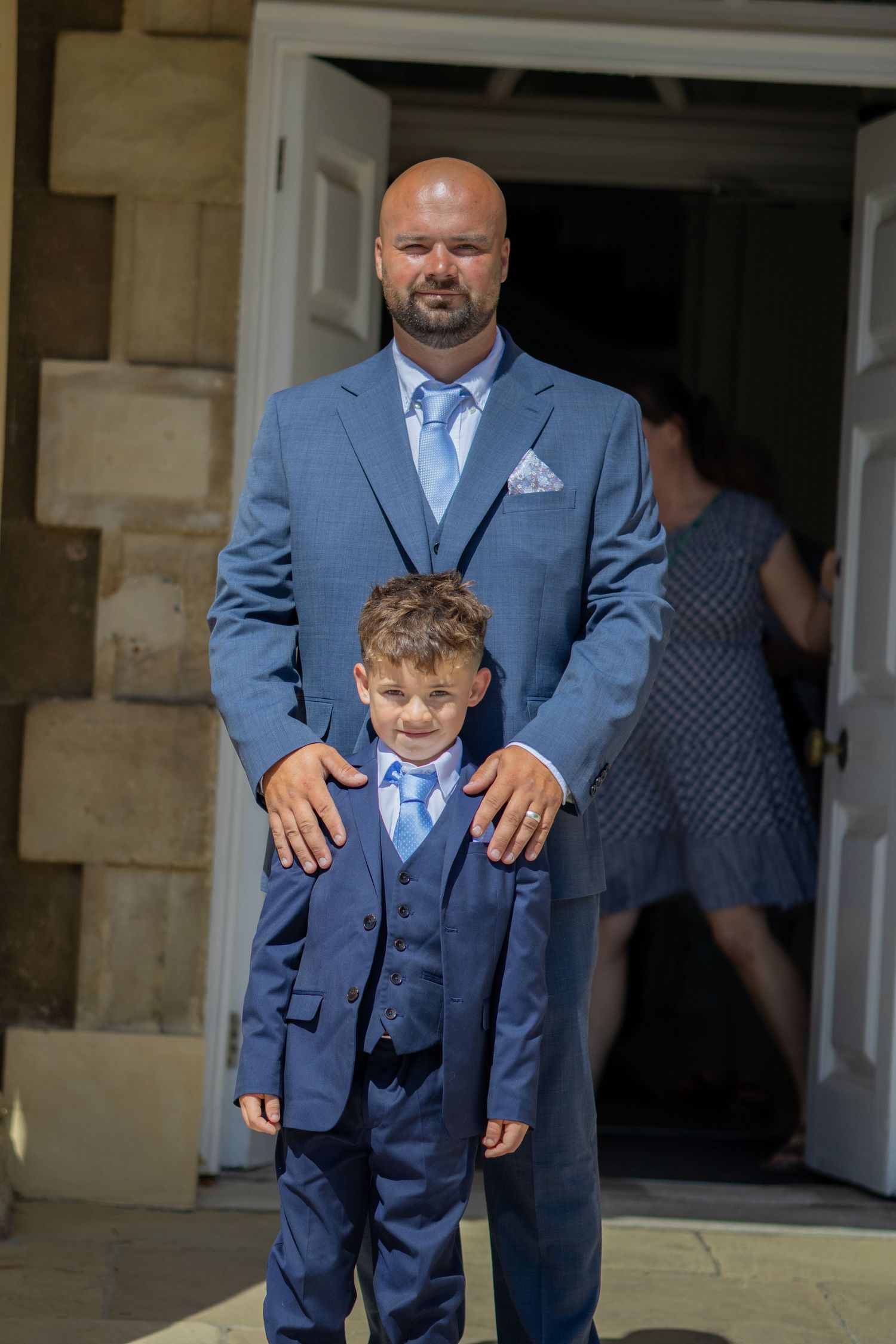 A bald groom in a blue suit stands proudly with a young boy in a matching blue suit outside a venue.
