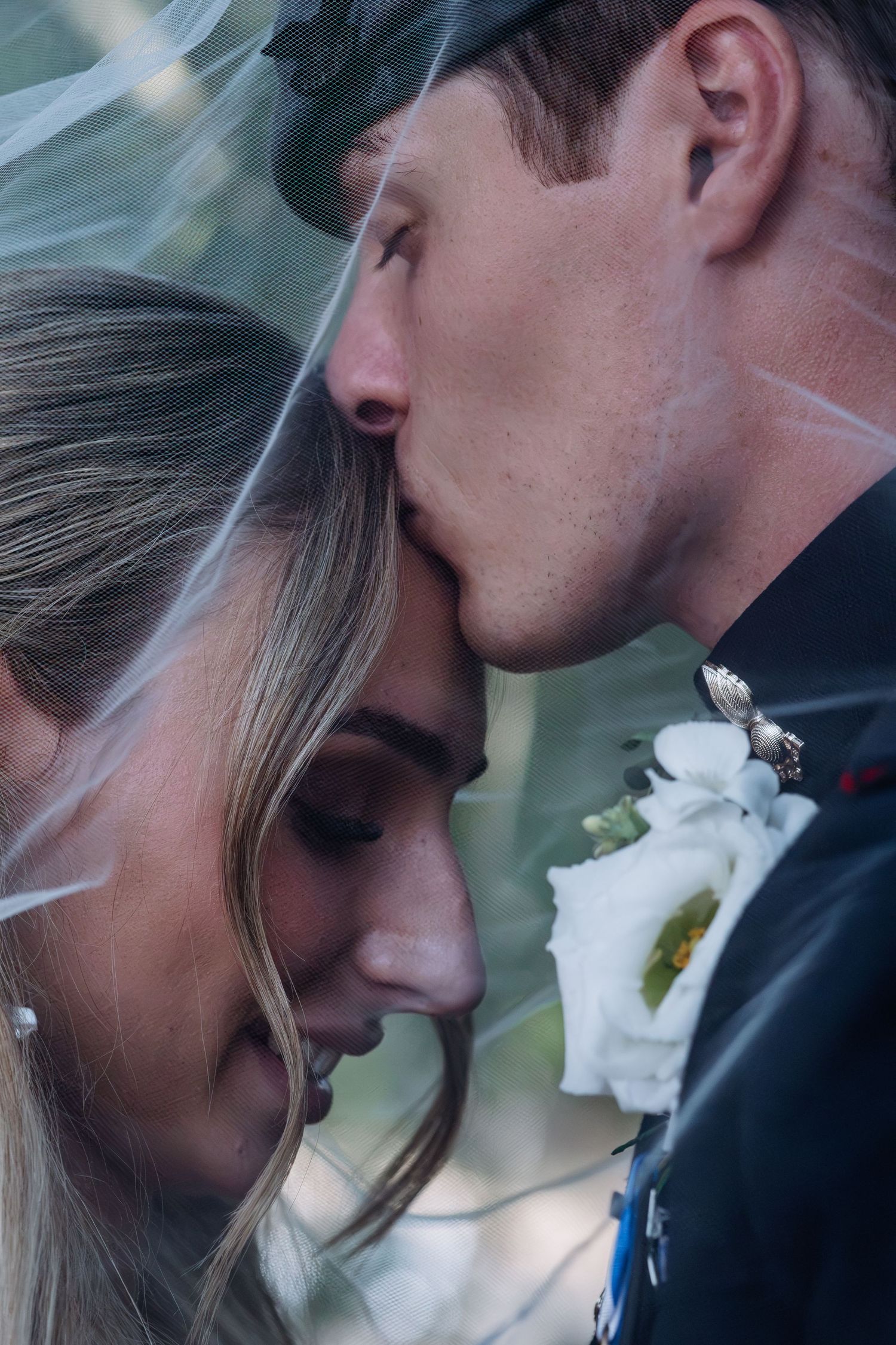 Groom in military uniform kisses bride on forehead under wedding veil.