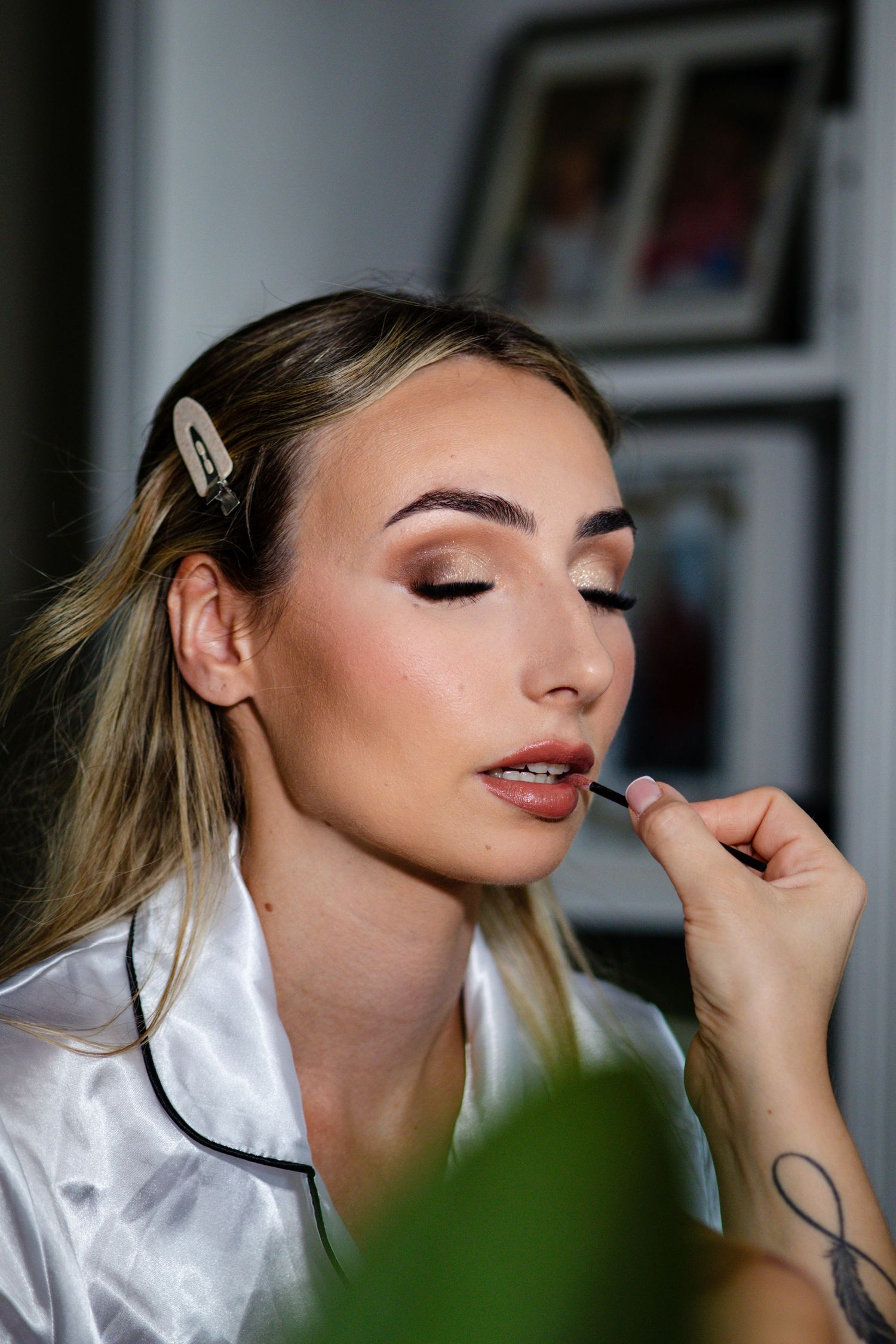 Makeup artist applying lipstick to a blonde woman in a white satin robe during bridal makeup session.