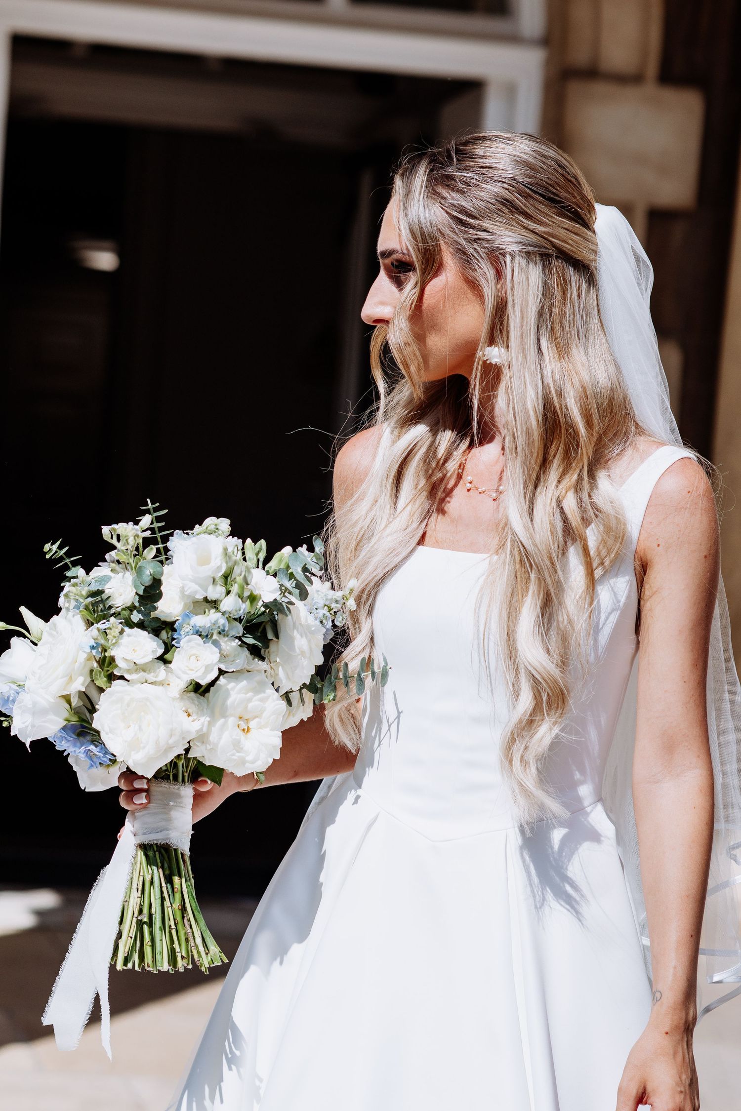 Elegant bride with long wavy blonde hair in white gown holds white and blue wedding bouquet.