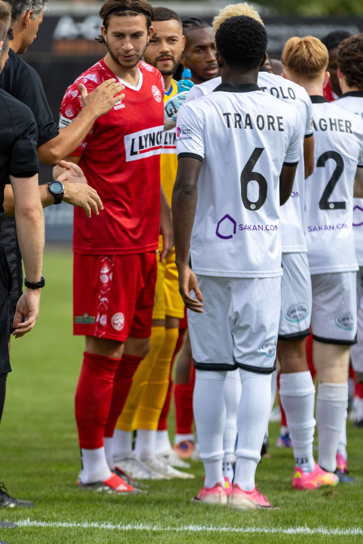 Football players in white Sakan.com kits including Traore 6 shaking hands with opponents in red.