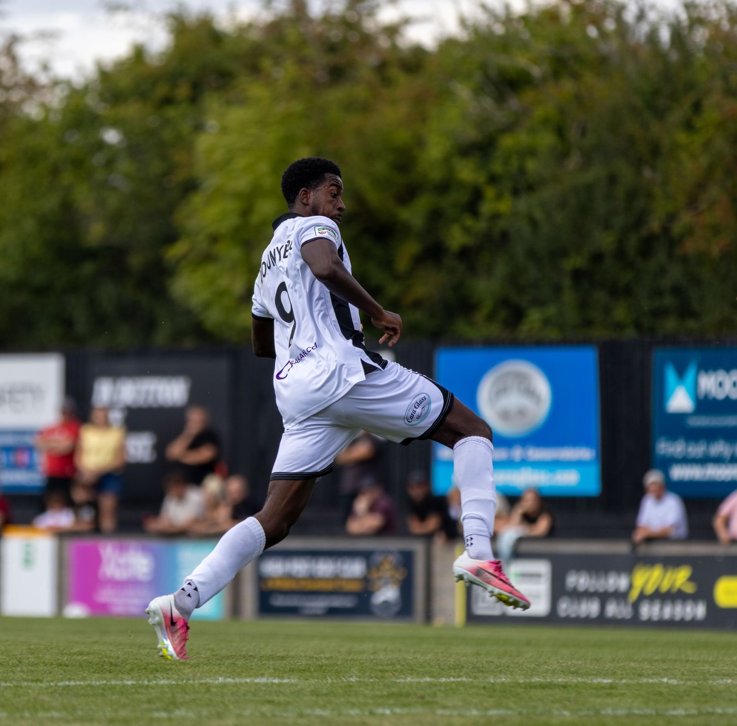 Football player number 9 in white kit jumping mid-air during an outdoor match with green trees in background.