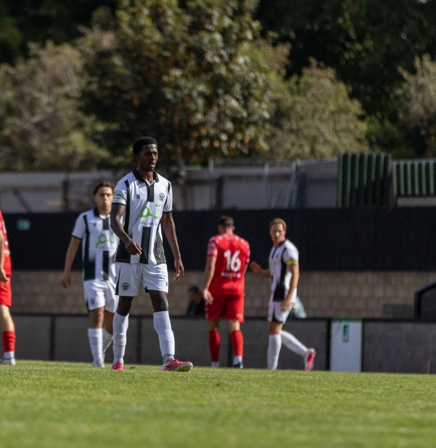 Footballer in black and white kit stands on pitch during match against red-kitted opponents.