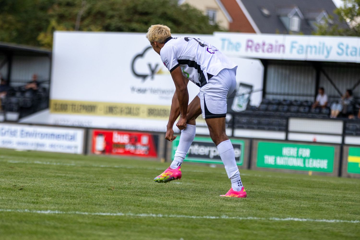 A footballer in white kit and pink boots bends forward near the Retain Family Stand during a match.