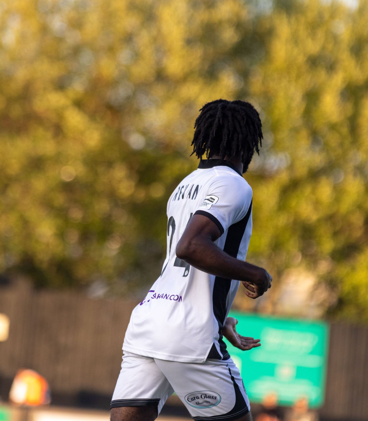 Football player in white and black strip jogging on pitch with trees in background.