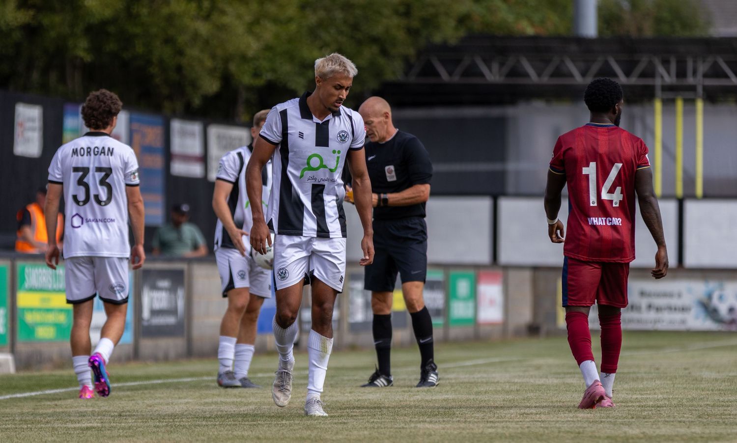 Football players in black and white striped kit during a match with referee and opposition in red.