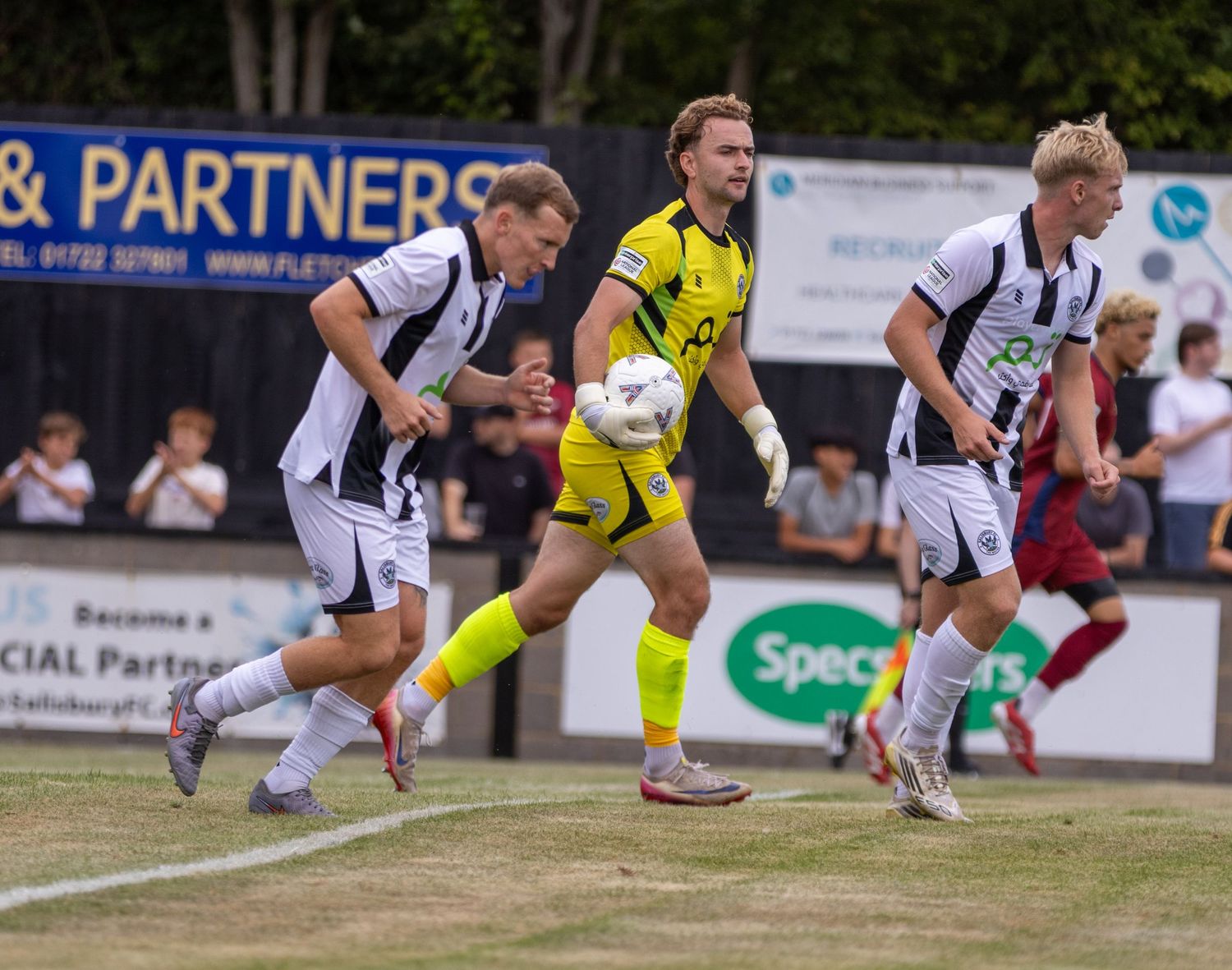 Non-league football players in black and white strip challenge a yellow goalkeeper holding the ball on a sunny day.