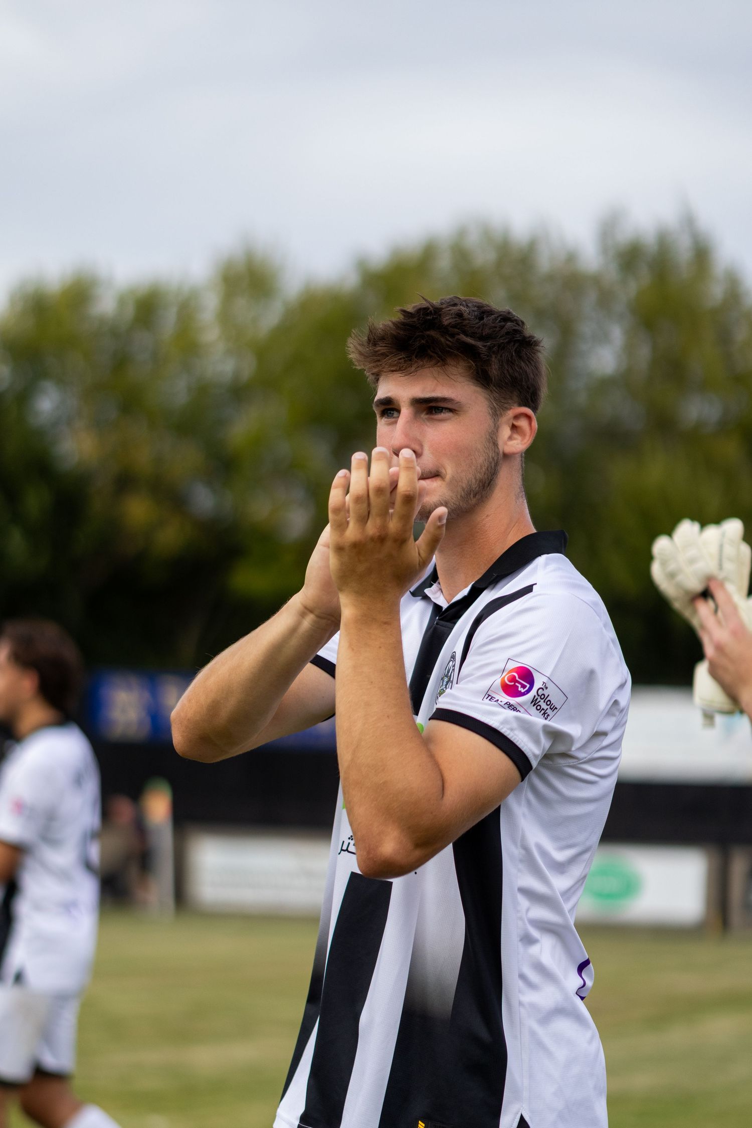 Young male footballer in black and white striped jersey applauds fans after a match on grass pitch.