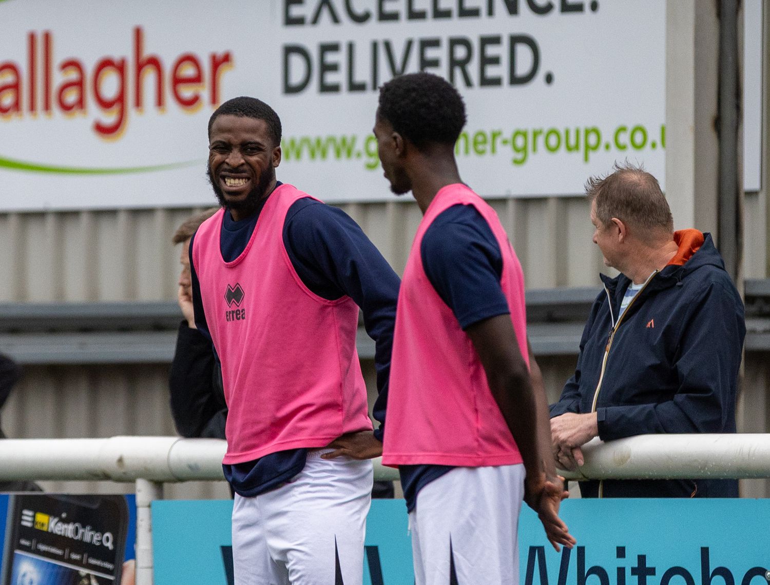 Two soccer players in pink bibs laugh on the sideline near advertising boards during a match.