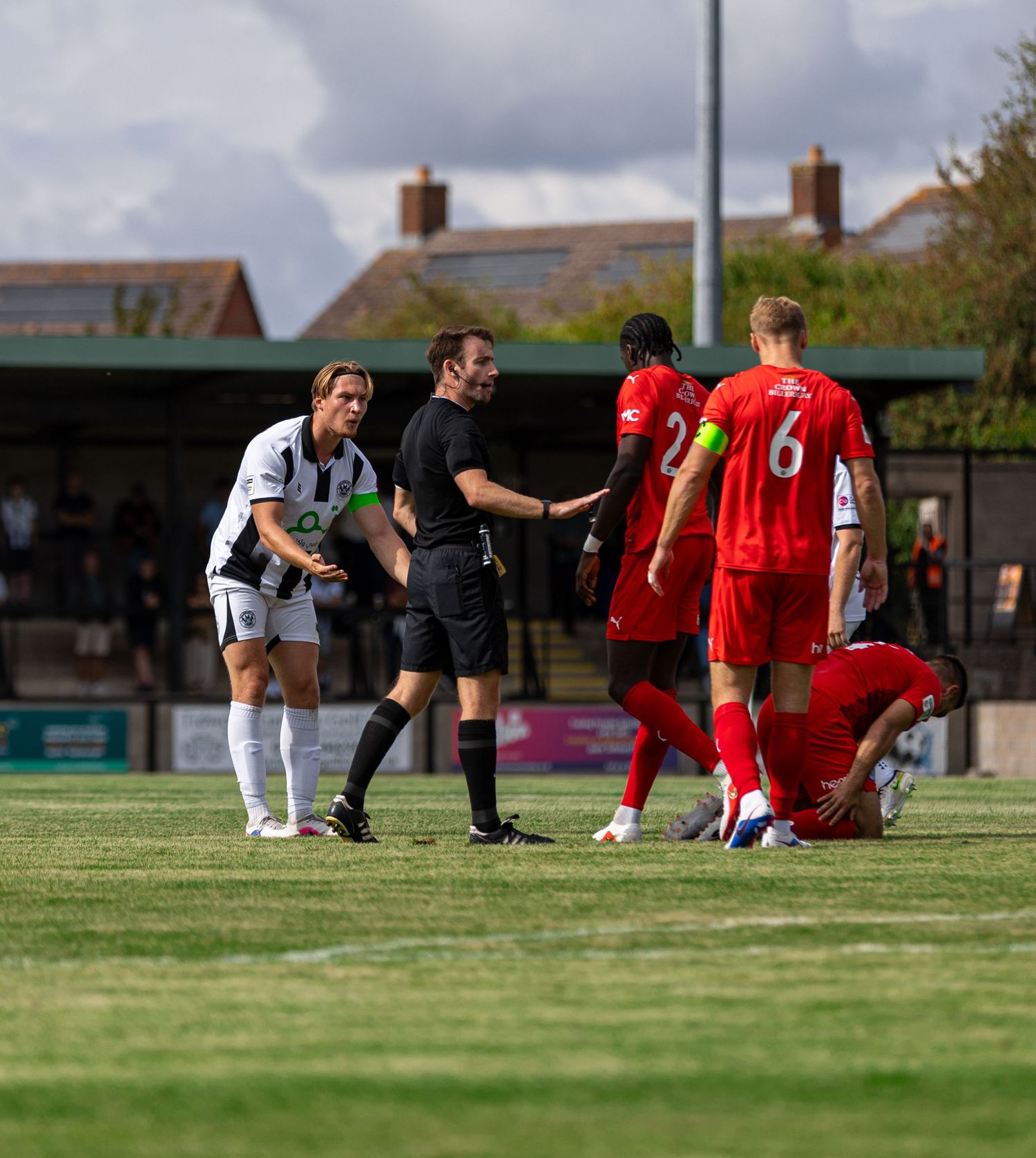 Football match scene with referee mediating between players in white and red kits on a sunny grass pitch.