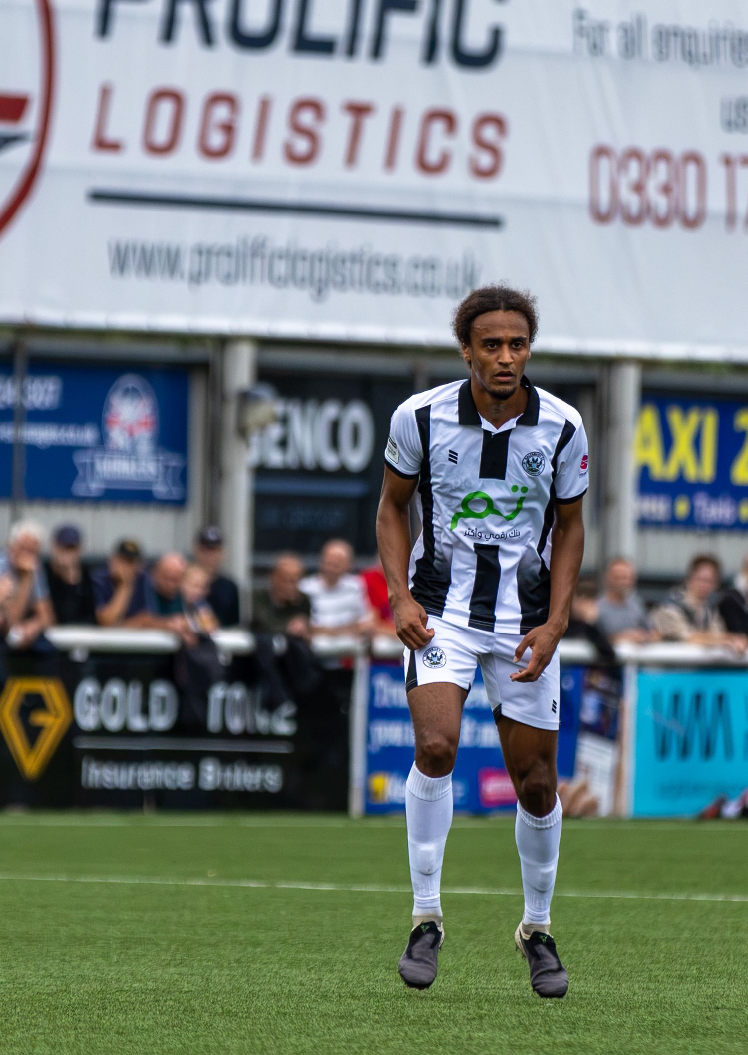 A footballer in black and white kit runs on the pitch during a match, with Prolific Logistics advertising boards behind.