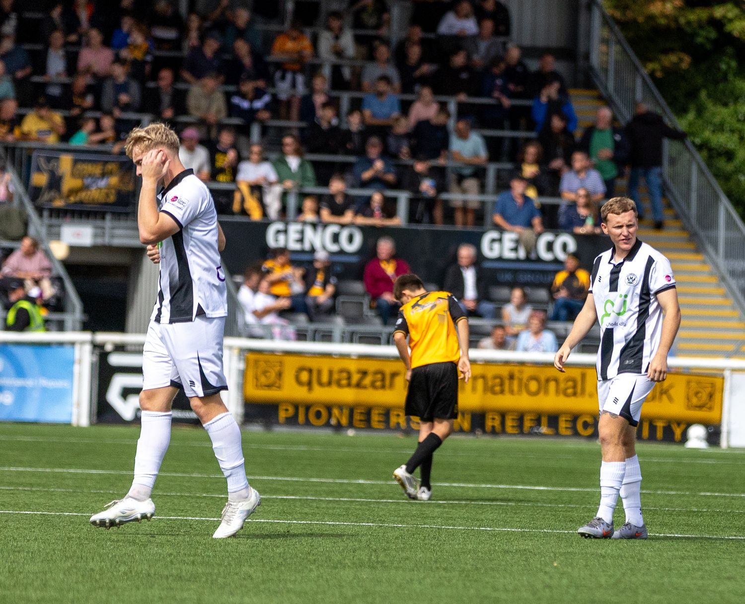 Football players in black and white kits react with frustration during a match, with a referee and crowd visible in background.